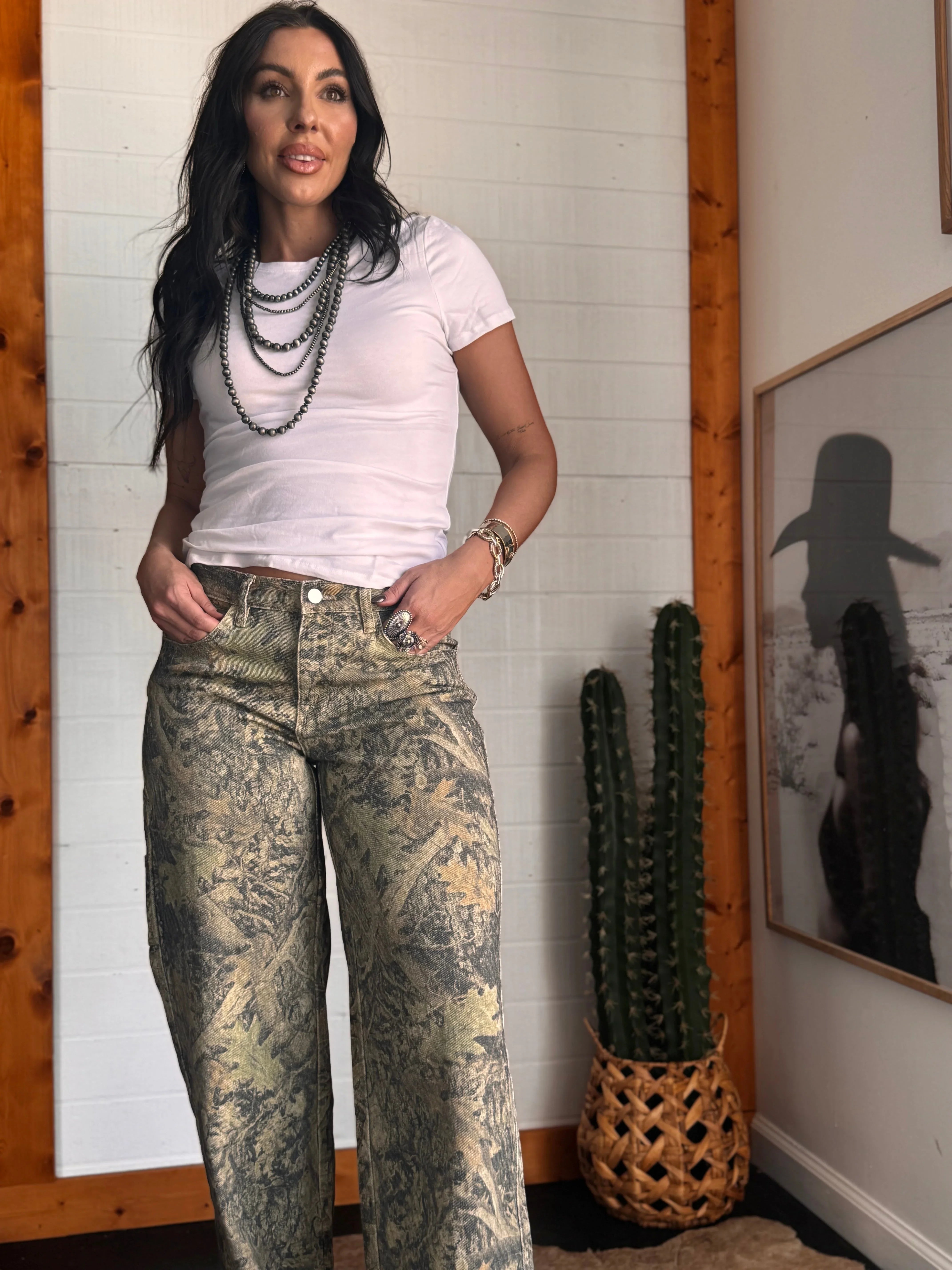 Woman wearing a white t-shirt and patterned jeans standing in a room with a cactus plant and framed picture on the wall.