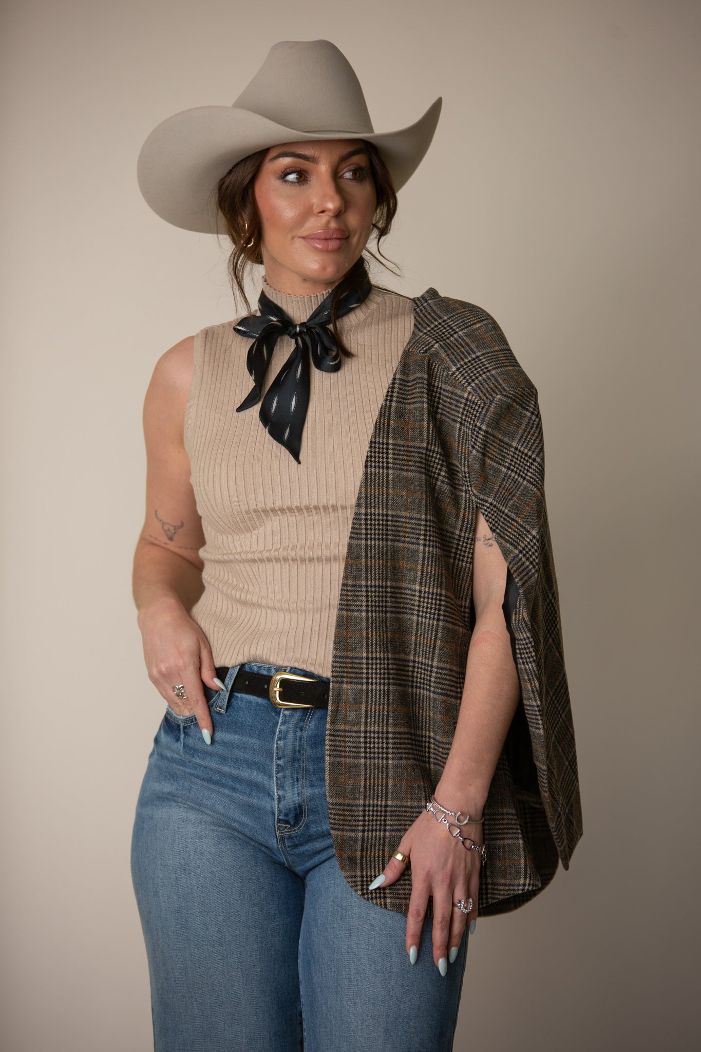 Woman in a beige cowboy hat, sleeveless ribbed top, plaid cape, jeans, and western scarf