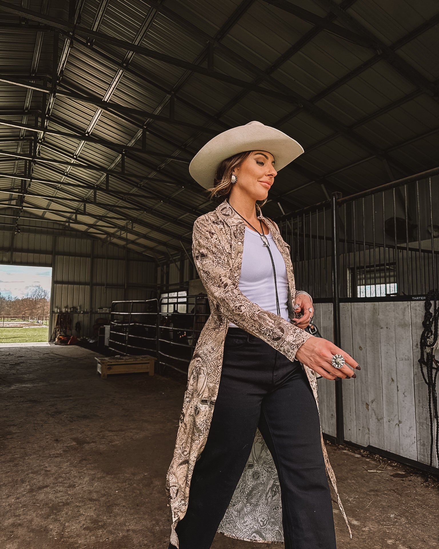Woman in western boho outfit with white cowboy hat, duster, and jewelry in a barn setting