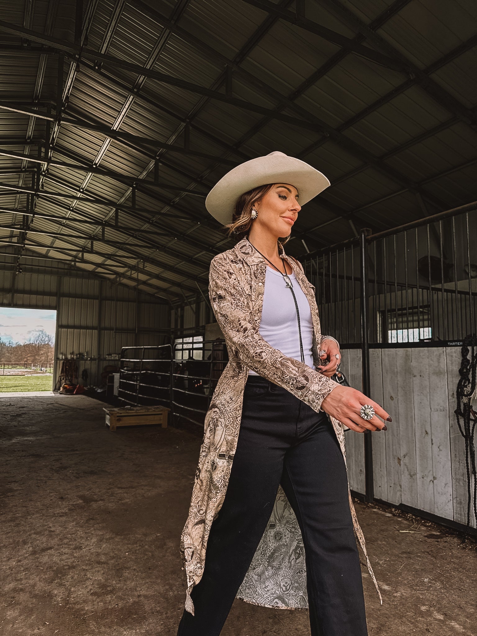 Woman in western boho outfit with white cowboy hat, duster, and jewelry in a barn setting