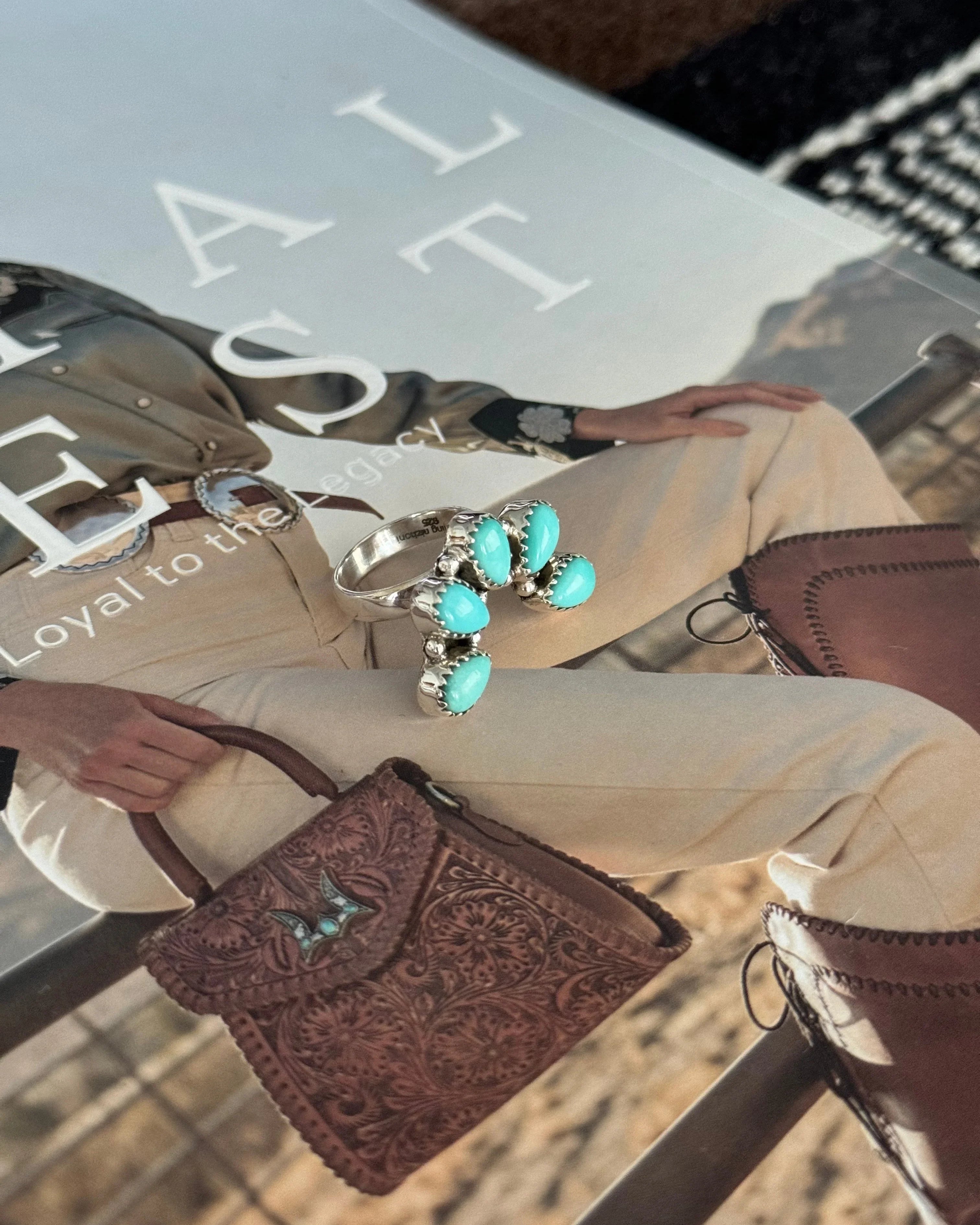 Turquoise western ring on magazine with cowgirl outfit, tooled leather bag, and cowboy boots.