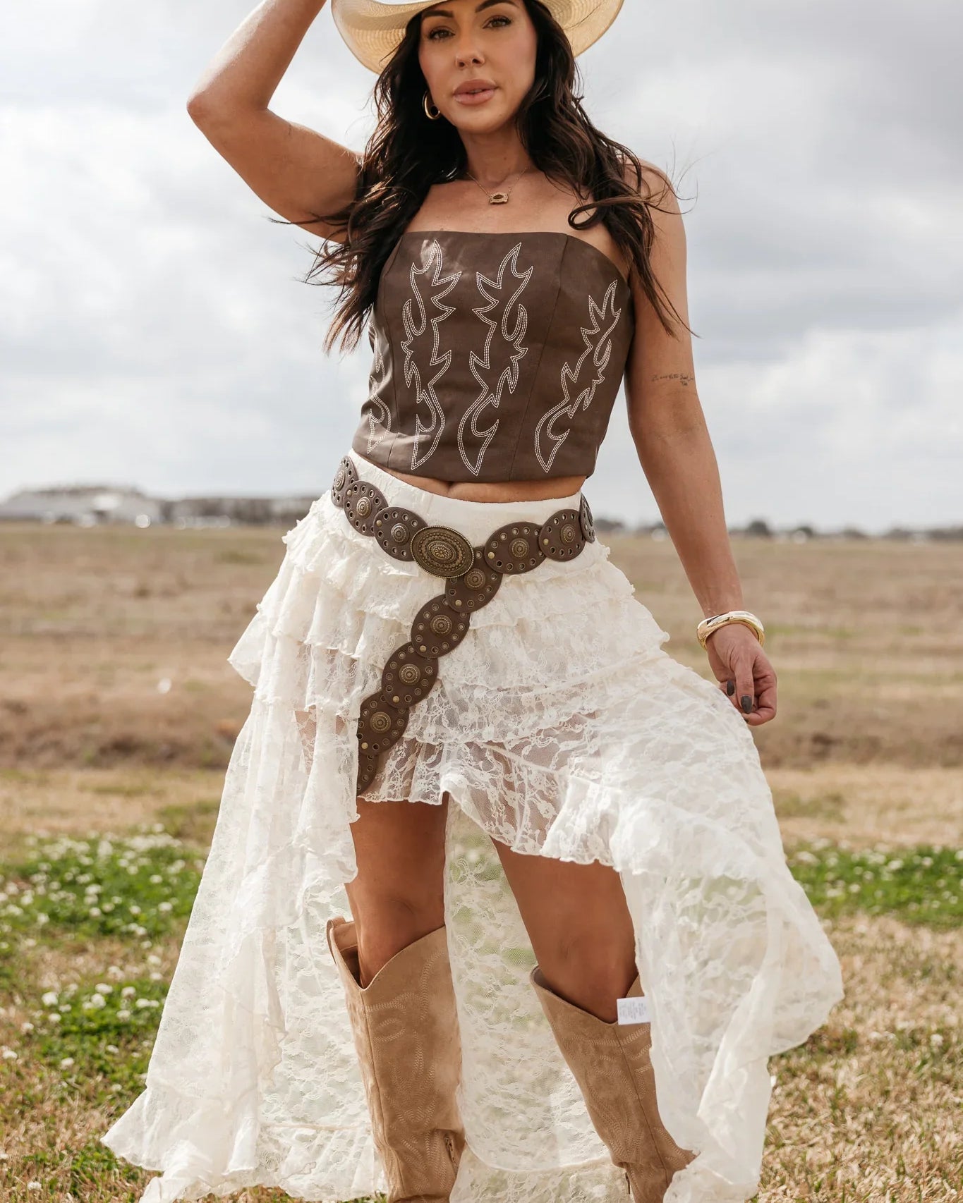Woman in western boho outfit with cowboy hat, brown corset top, lace skirt, and boots outdoors