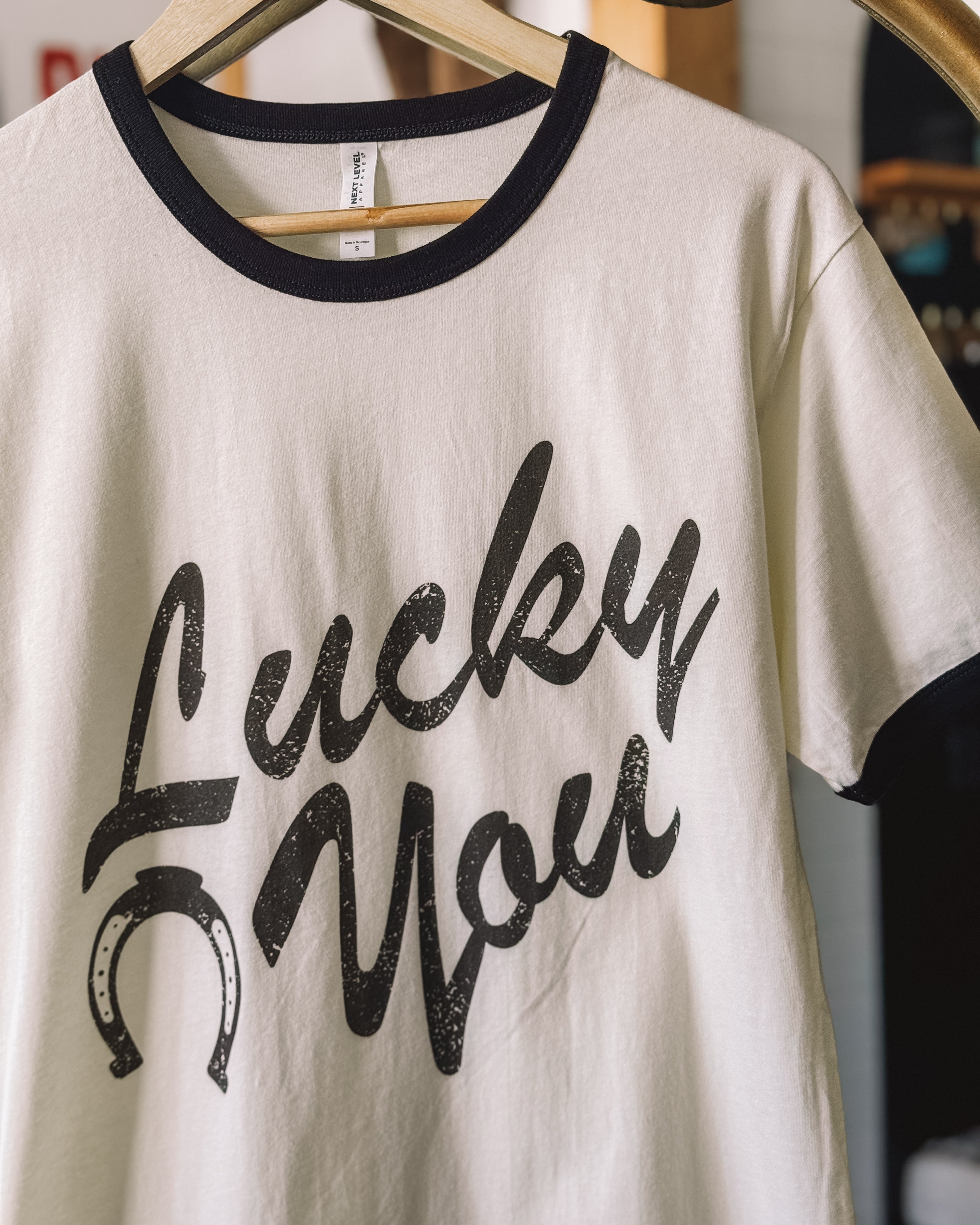 Western graphic tee with 'Lucky You' text and horseshoe, white with black trim, hanging on hanger