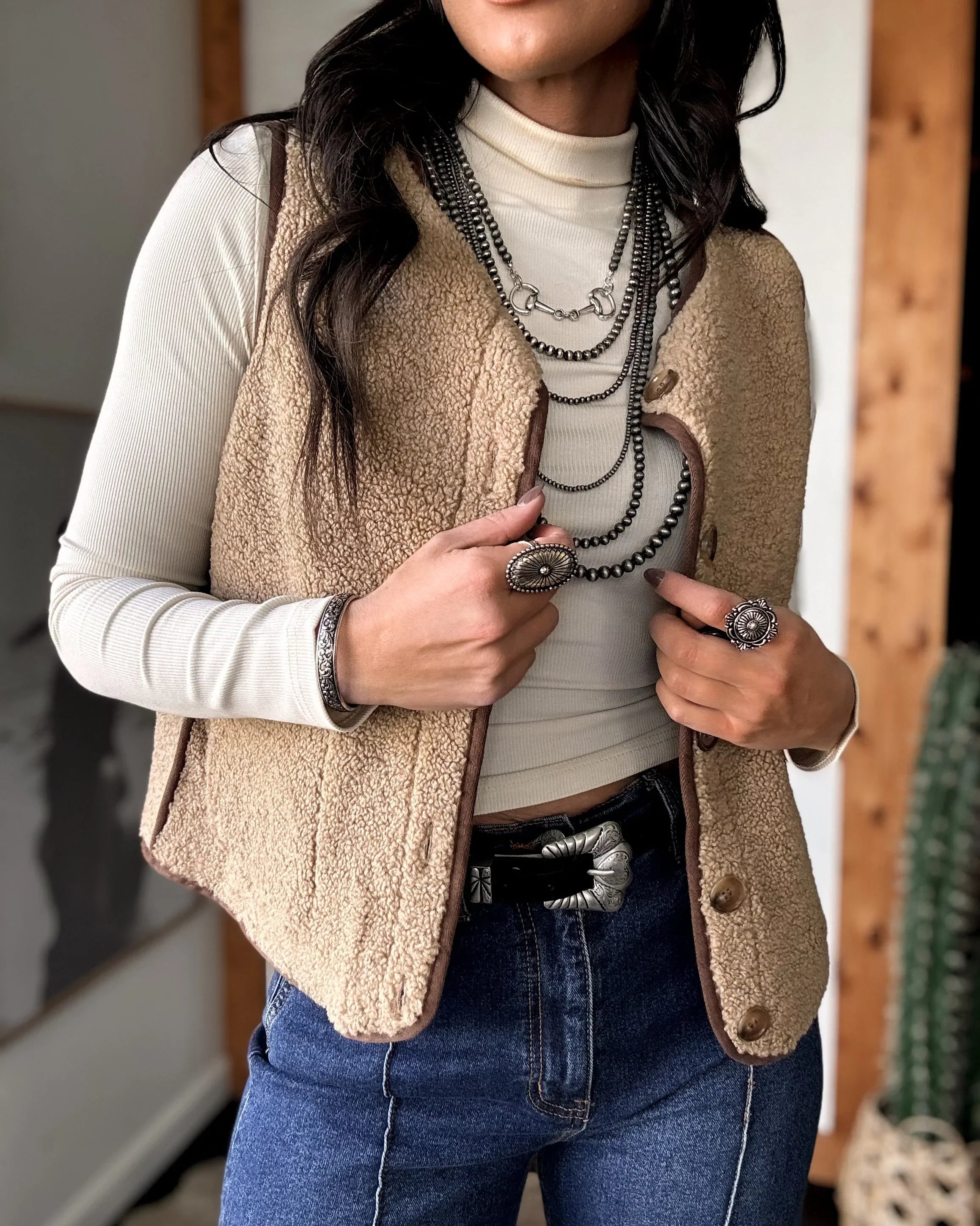 Woman wearing a beige vest over a white shirt with blue jeans indoors.