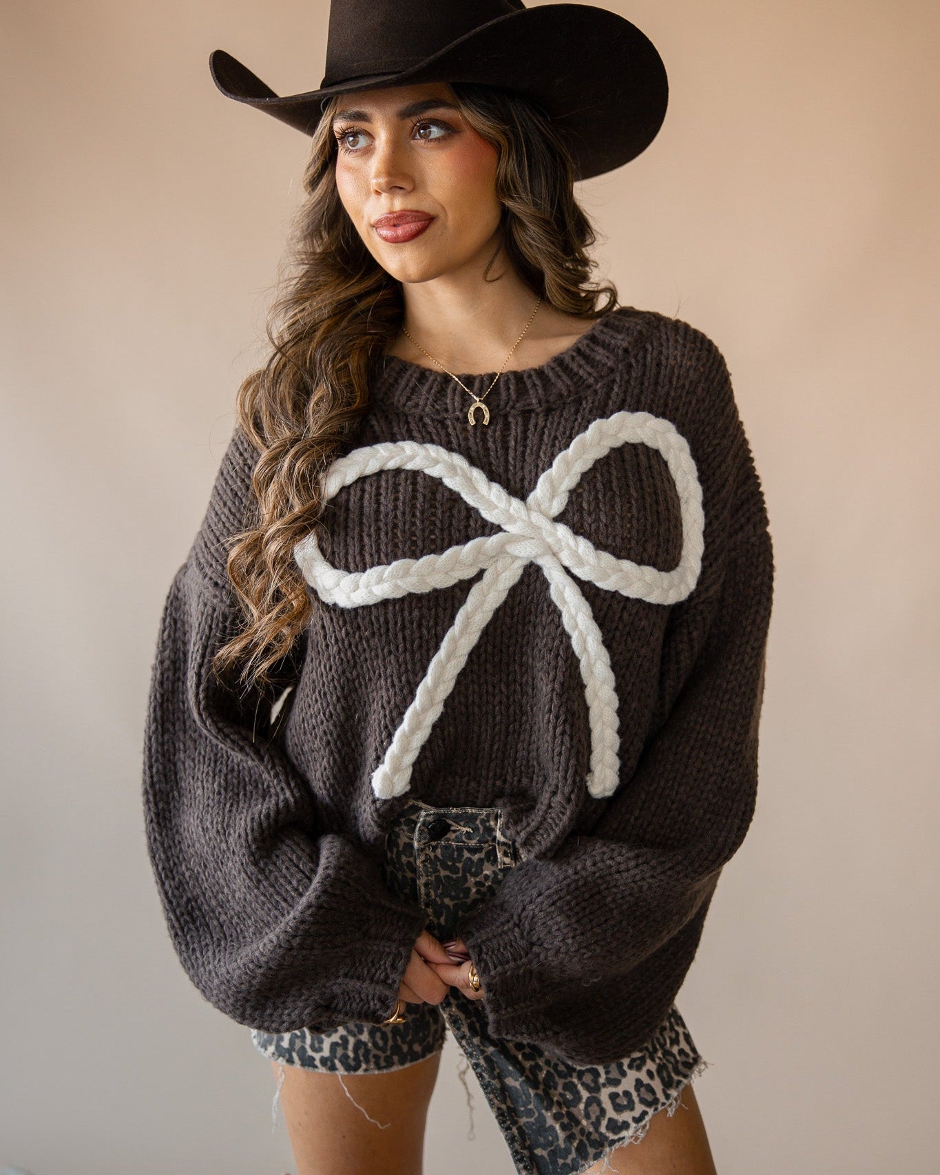 Woman in western outfit with black cowboy hat, chunky brown sweater with white bow, and leopard print shorts