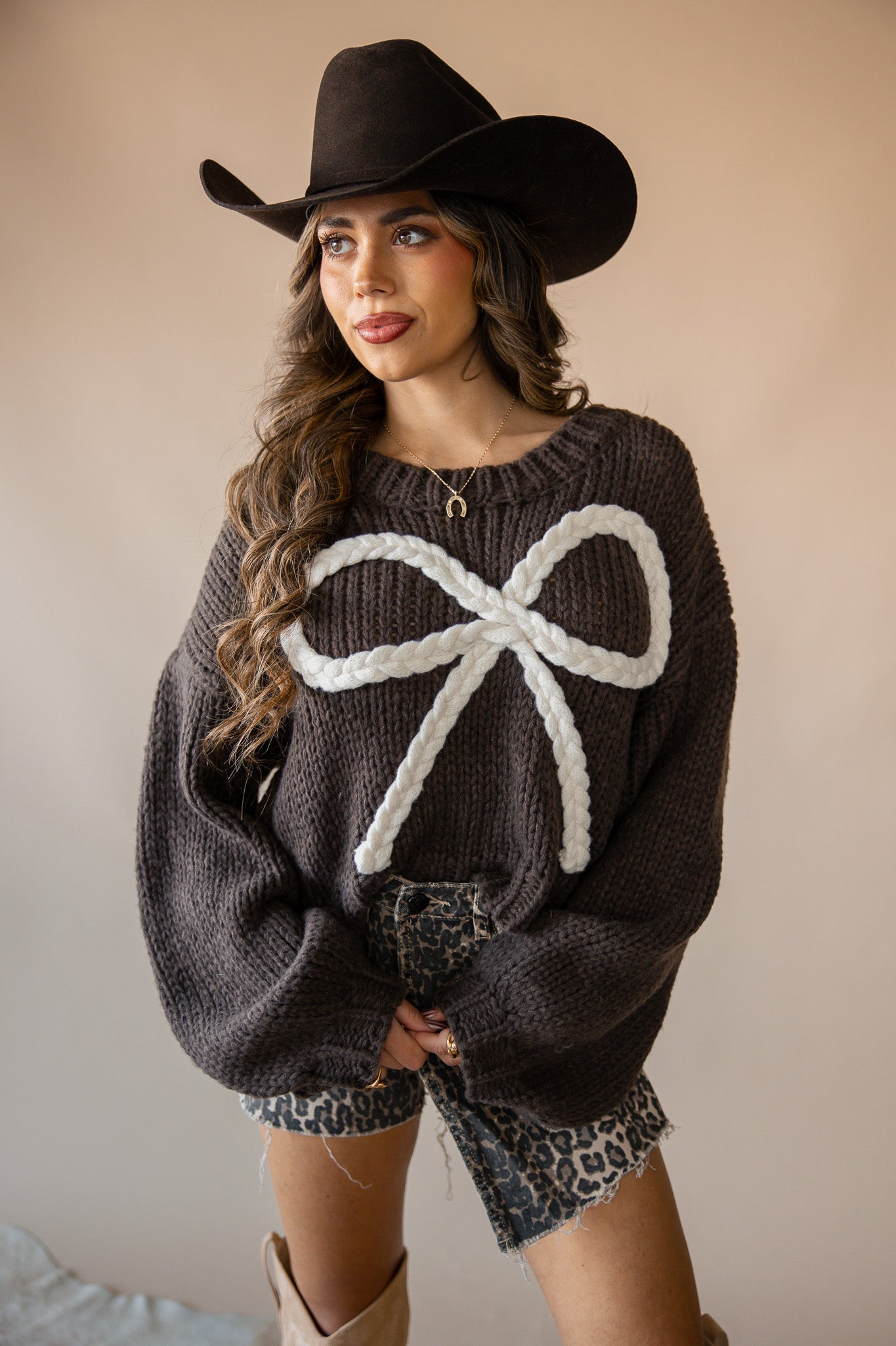 Woman in western outfit with black cowboy hat, chunky brown sweater with white bow, and leopard print shorts