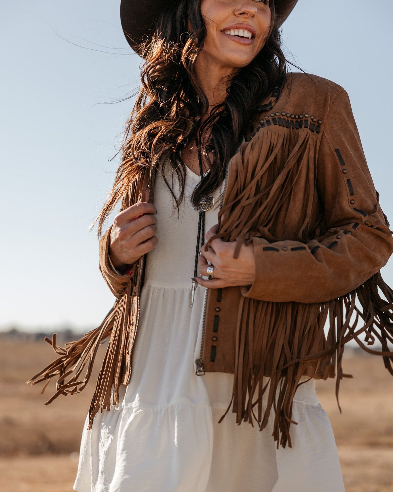 Woman in brown suede fringe jacket, white boho dress, and black cowboy hat outdoors
