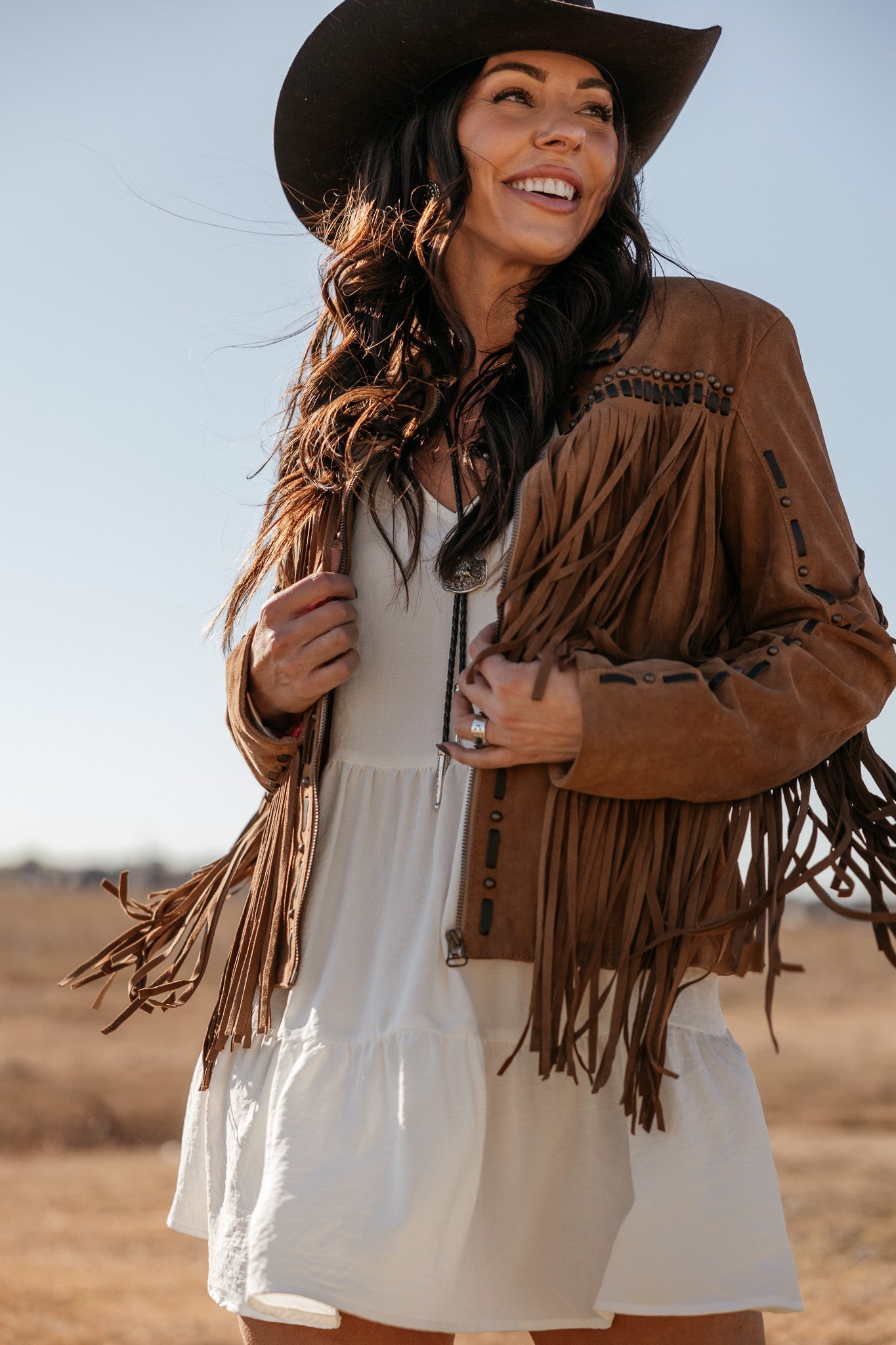 Woman in brown suede fringe jacket, white boho dress, and black cowboy hat outdoors