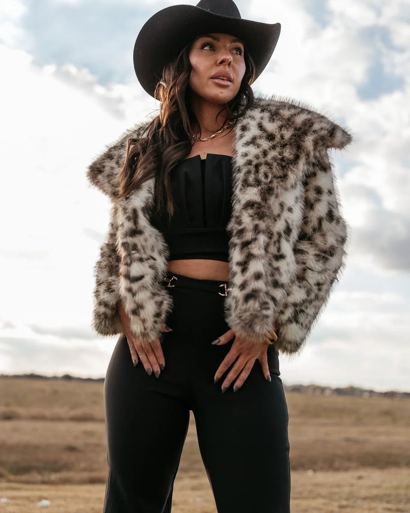 Woman in black cowgirl hat, faux fur animal print jacket, and western boho outfit outdoors