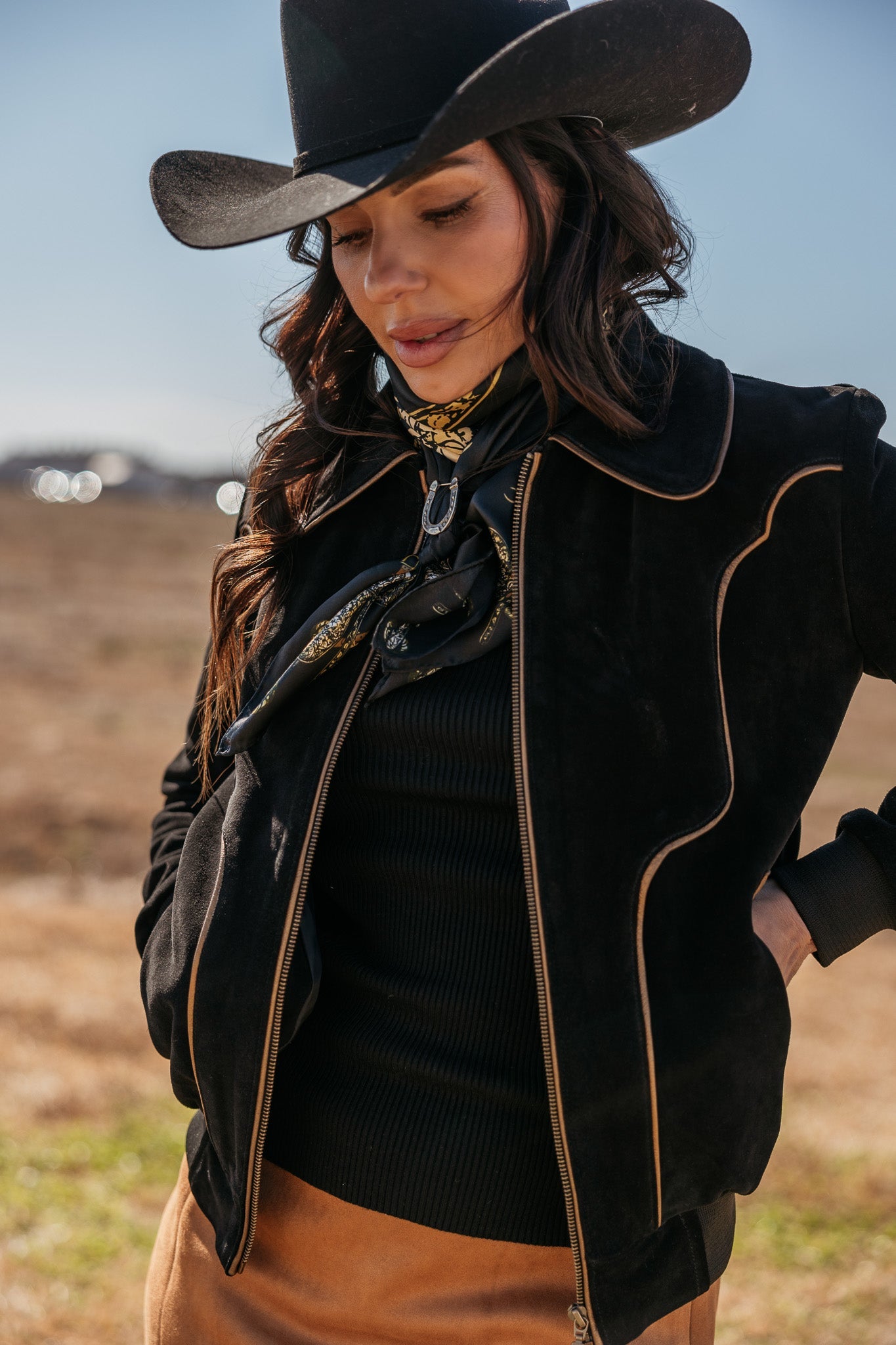 Woman in black cowboy hat and western jacket with scarf, standing outdoors in boho style.