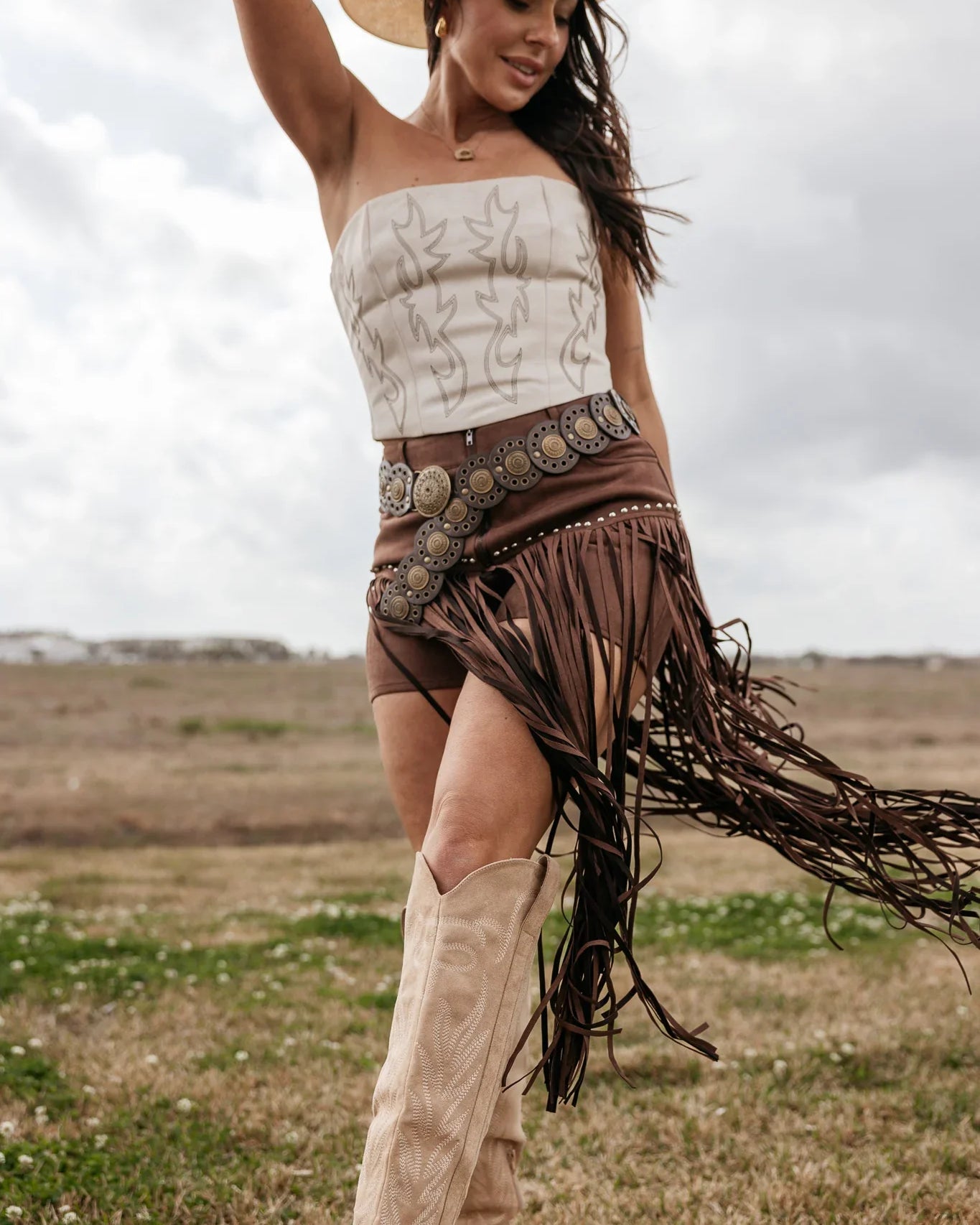 Woman in cowgirl hat, western boots, fringe skirt, and strapless top posing outdoors
