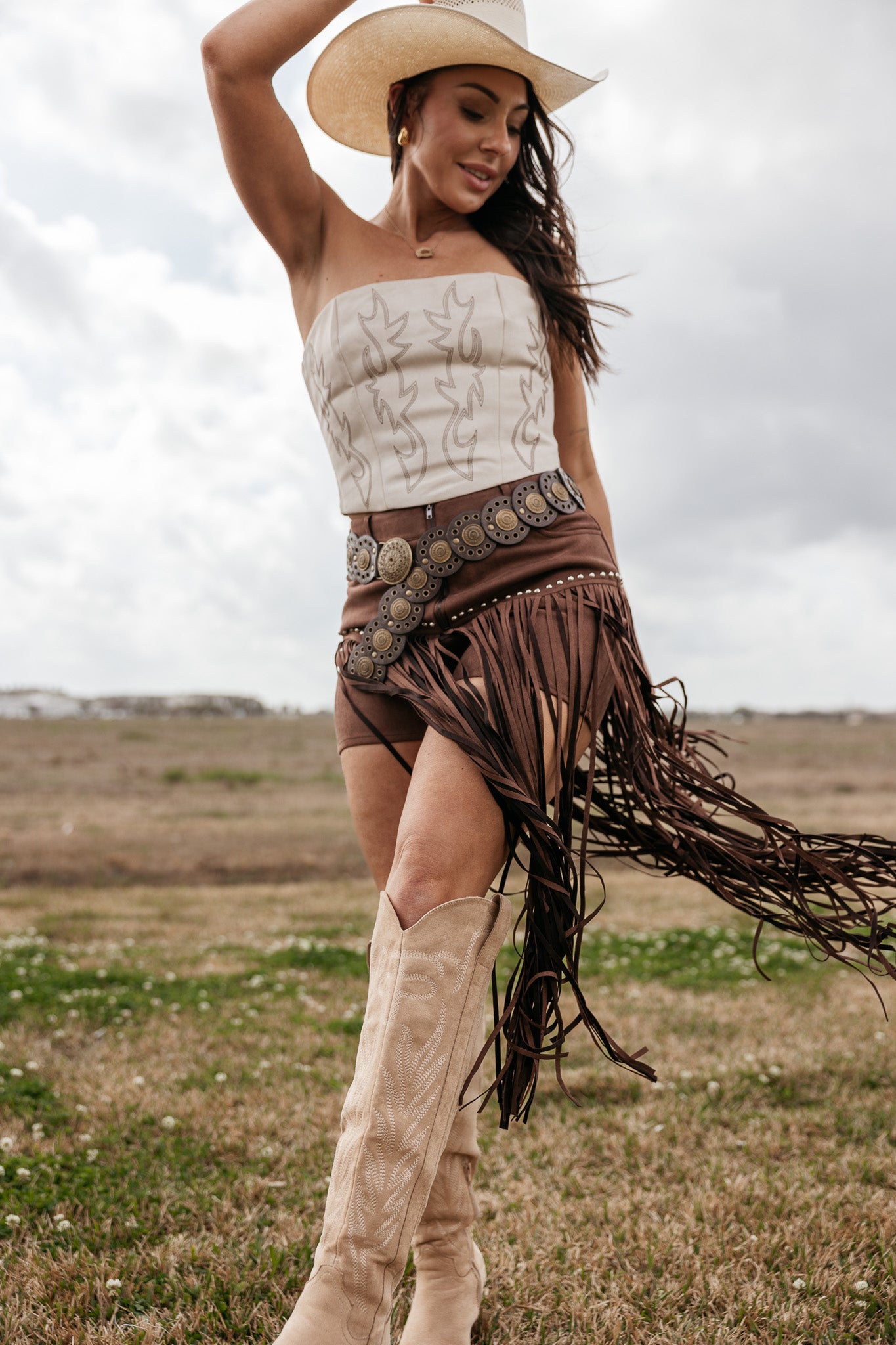 Woman in cowgirl hat, western boots, fringe skirt, and strapless top posing outdoors