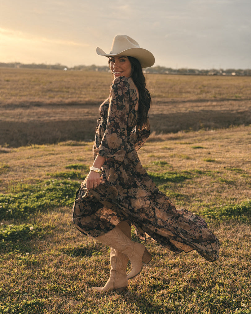 Woman in floral western dress, cowboy hat, and boots standing in a field at sunset