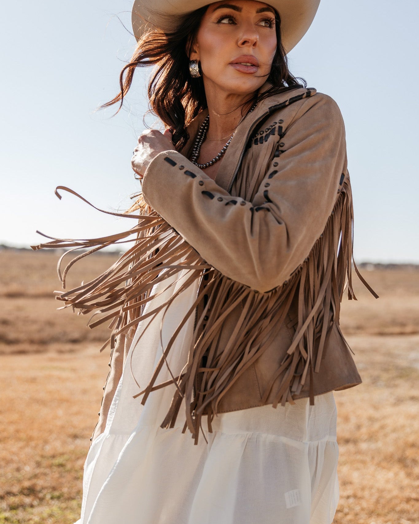 Woman in a fringed suede jacket, white western dress, and cowboy hat outdoors