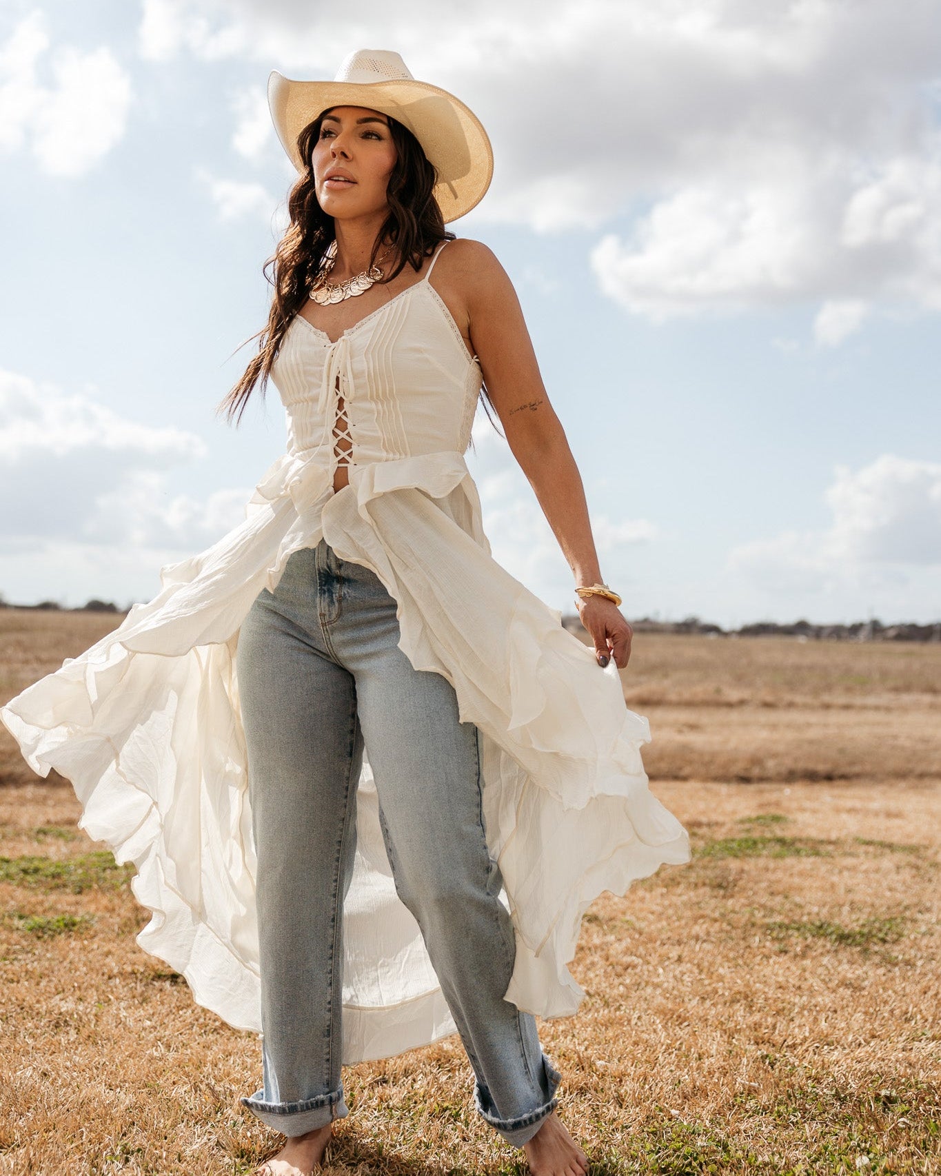 Woman in a white western lace-up top, cowboy hat, and blue jeans standing barefoot outdoors