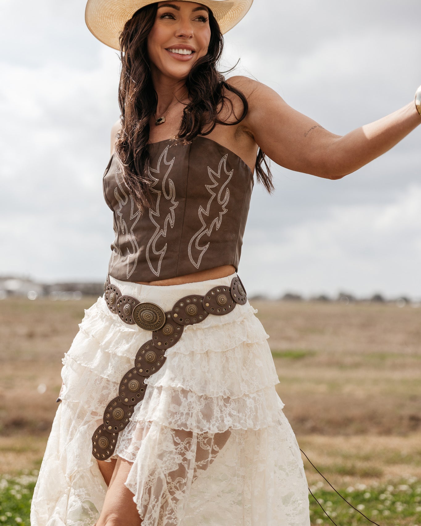 Woman in western boho outfit with cowboy hat, brown corset, lace skirt, and concho belt outdoors
