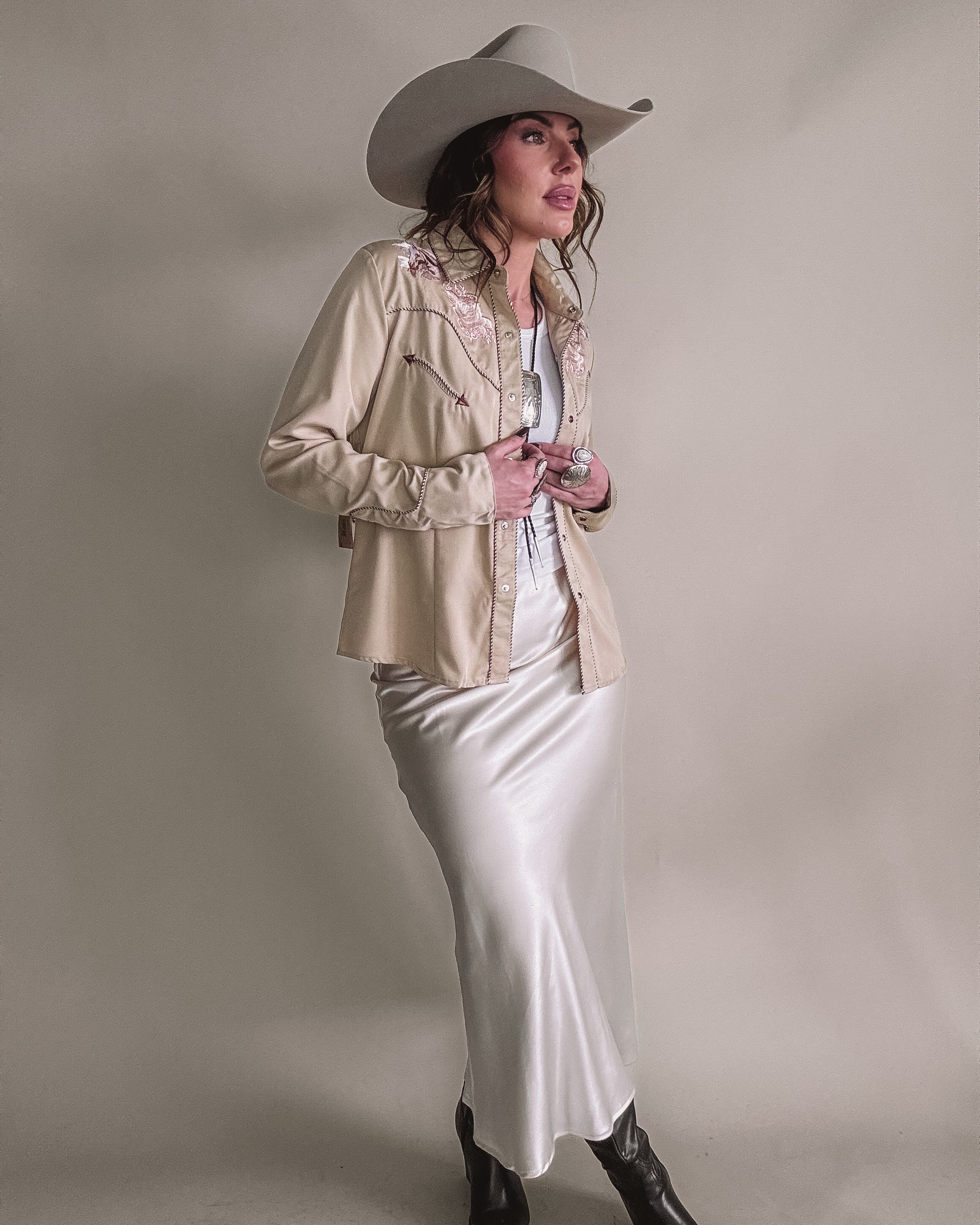 Woman in western cowgirl outfit with beige embroidered jacket, satin skirt, cowboy hat, and boots