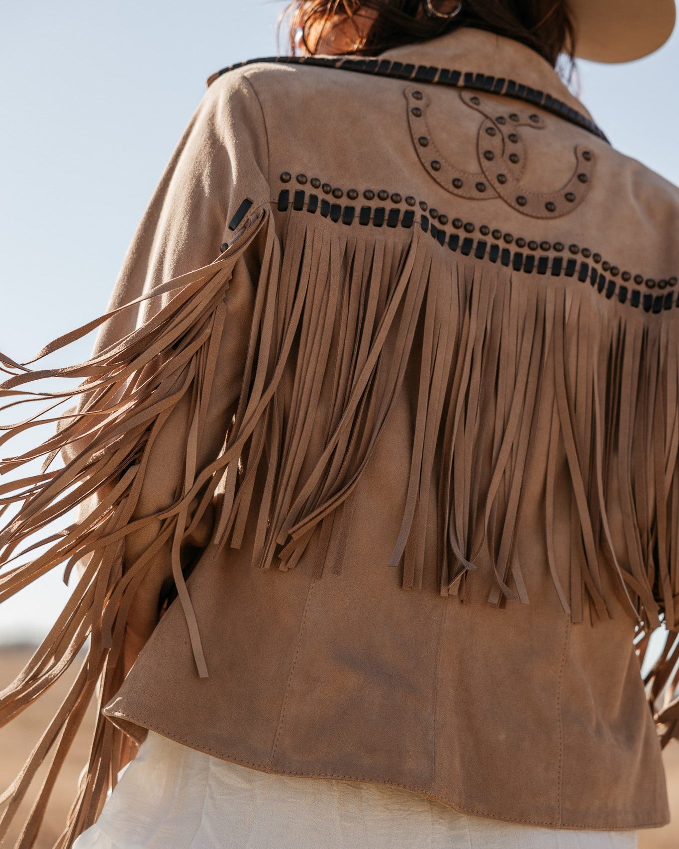 Woman wearing tan suede fringe jacket, western style, outdoors with cowboy hat