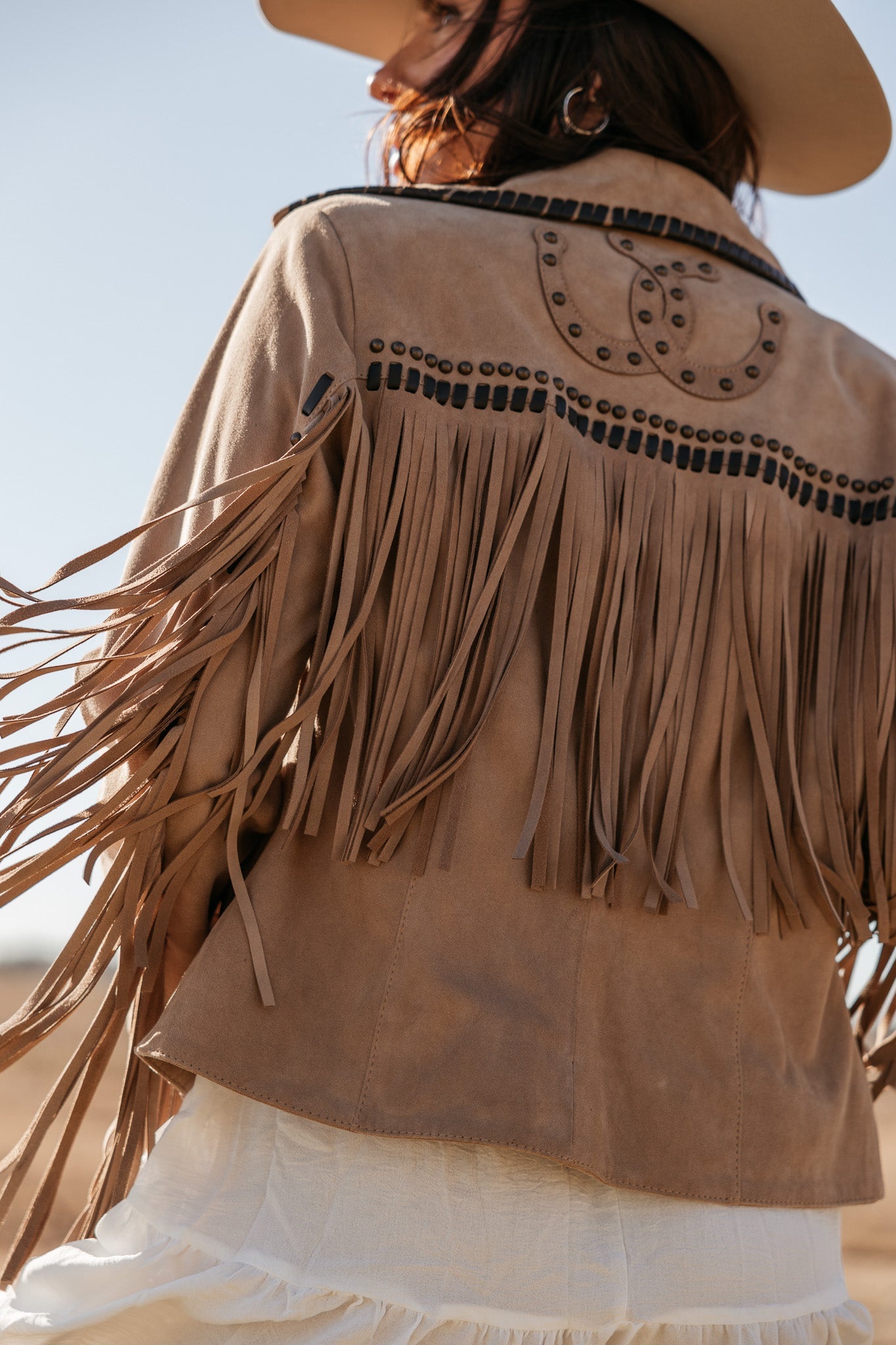 Woman wearing tan suede fringe jacket, western style, outdoors with cowboy hat