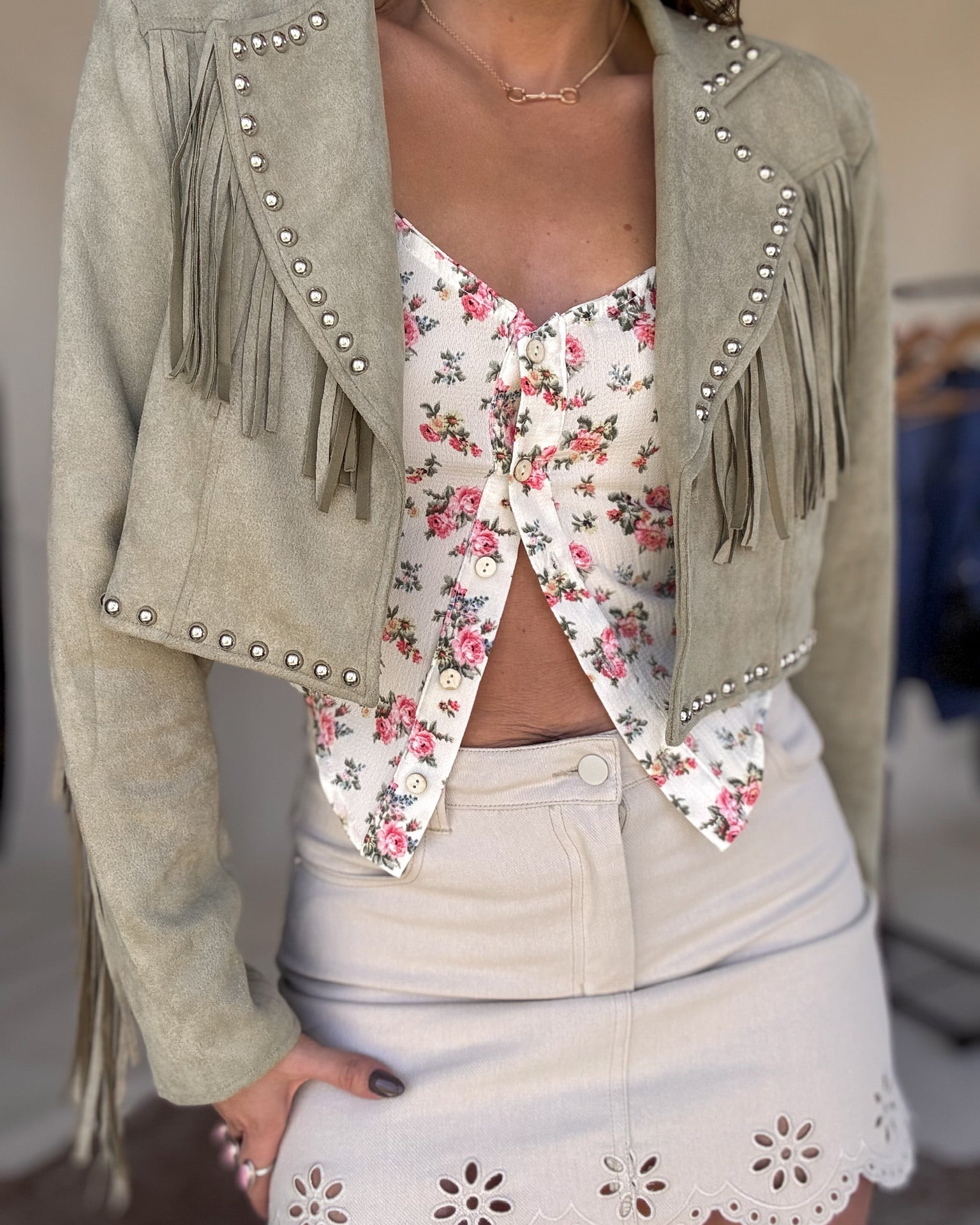 Western boho outfit with sage fringe jacket, floral top, and beige eyelet mini skirt
