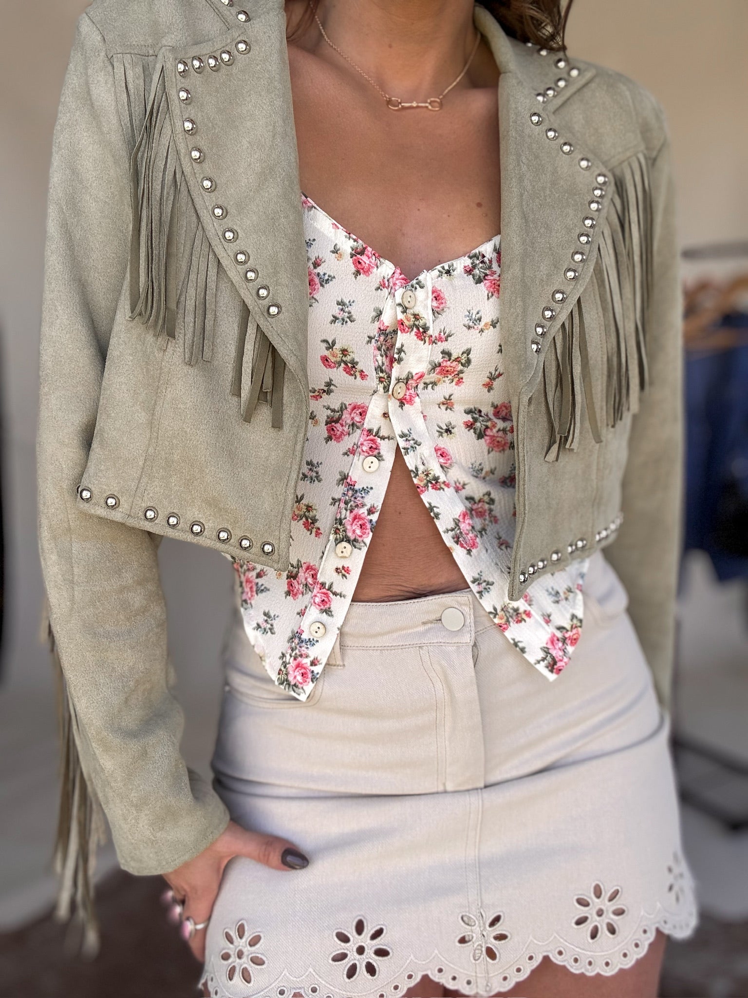 Western boho outfit with sage fringe jacket, floral top, and beige eyelet mini skirt