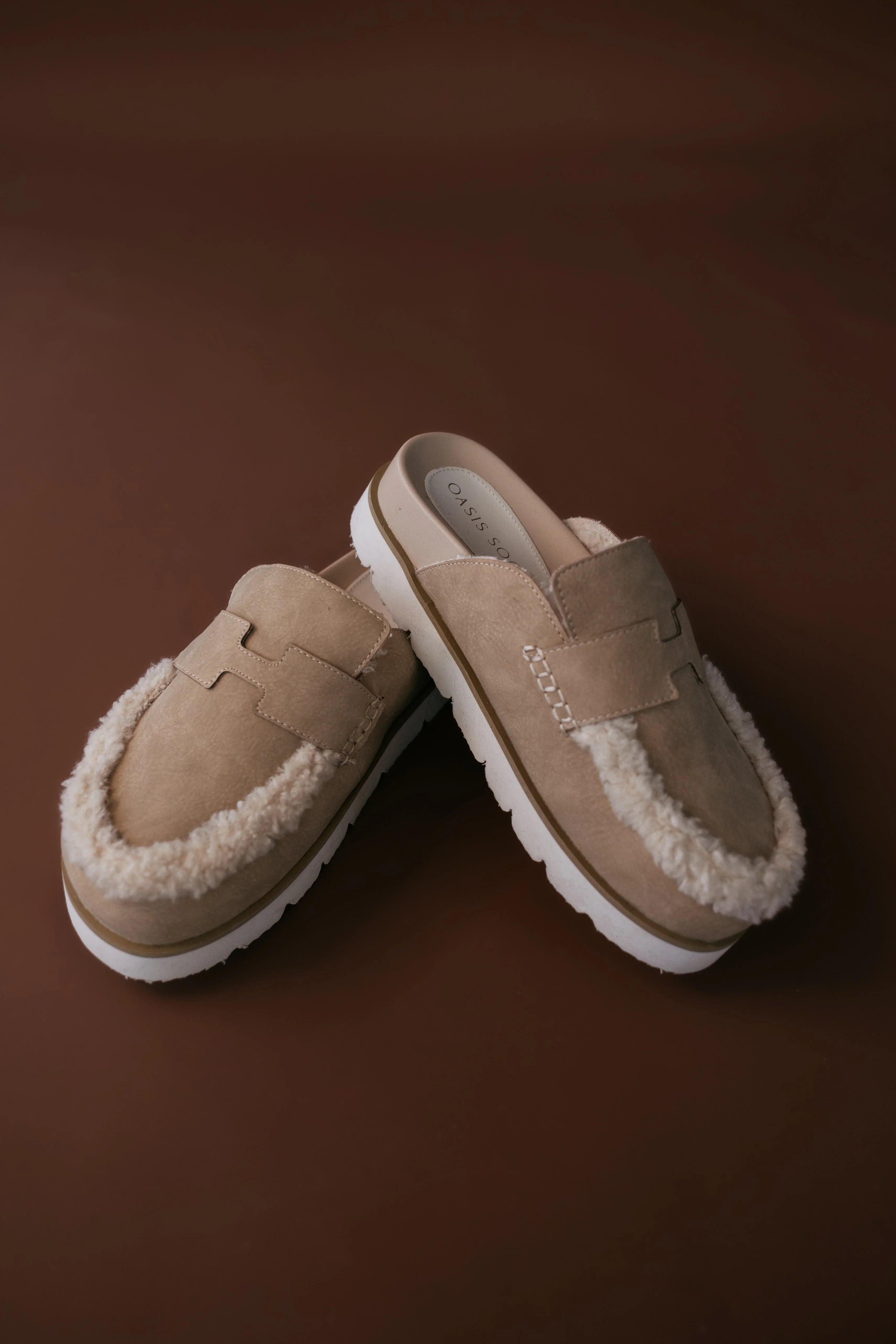 Pair of beige slippers with white fur lining on a brown background