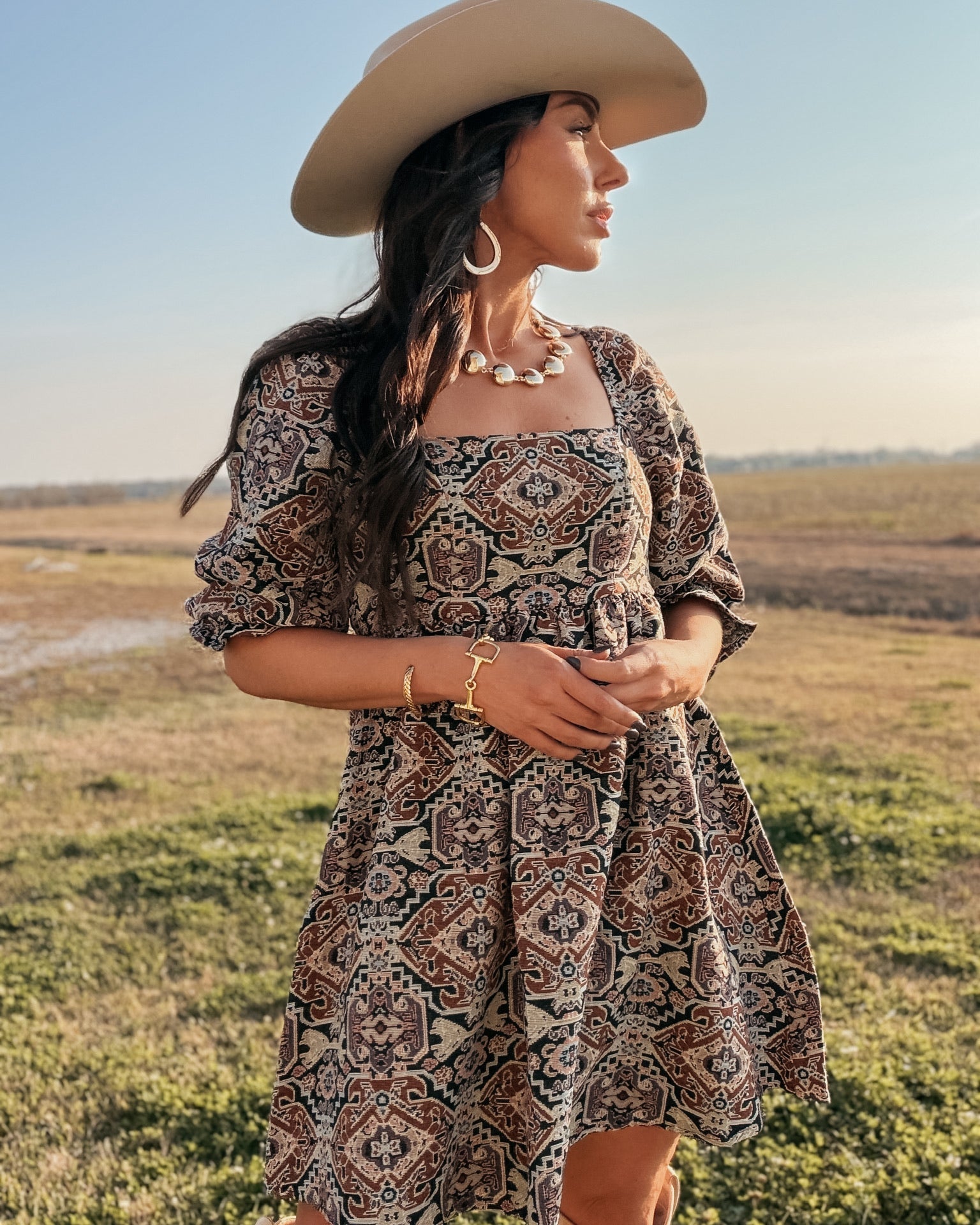 Woman in western boho print dress, cowboy hat, and boots standing outdoors at sunset.