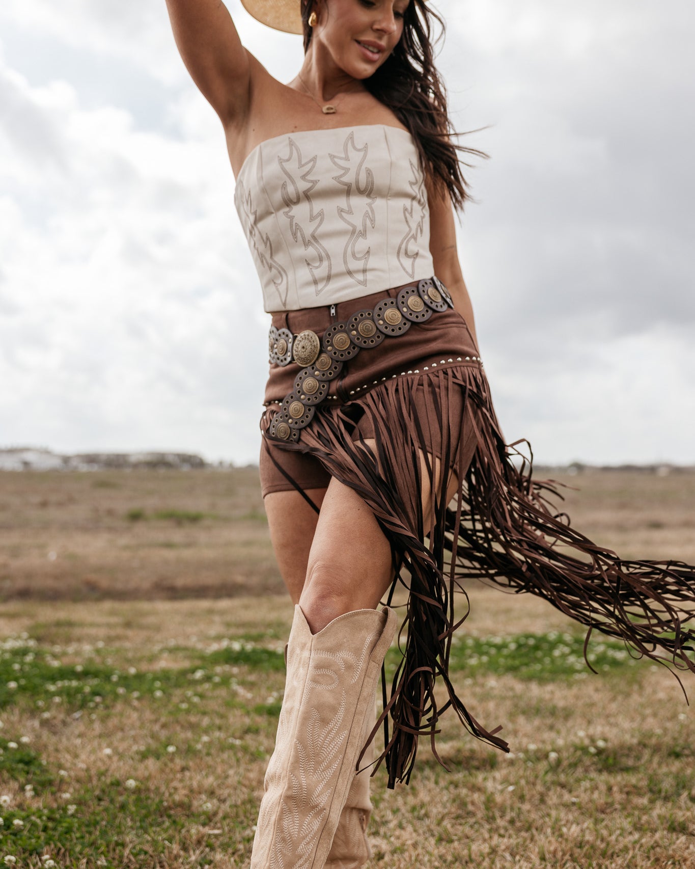 Woman in a cowboy outfit with a hat, top, and fringed skirt standing in an open field.