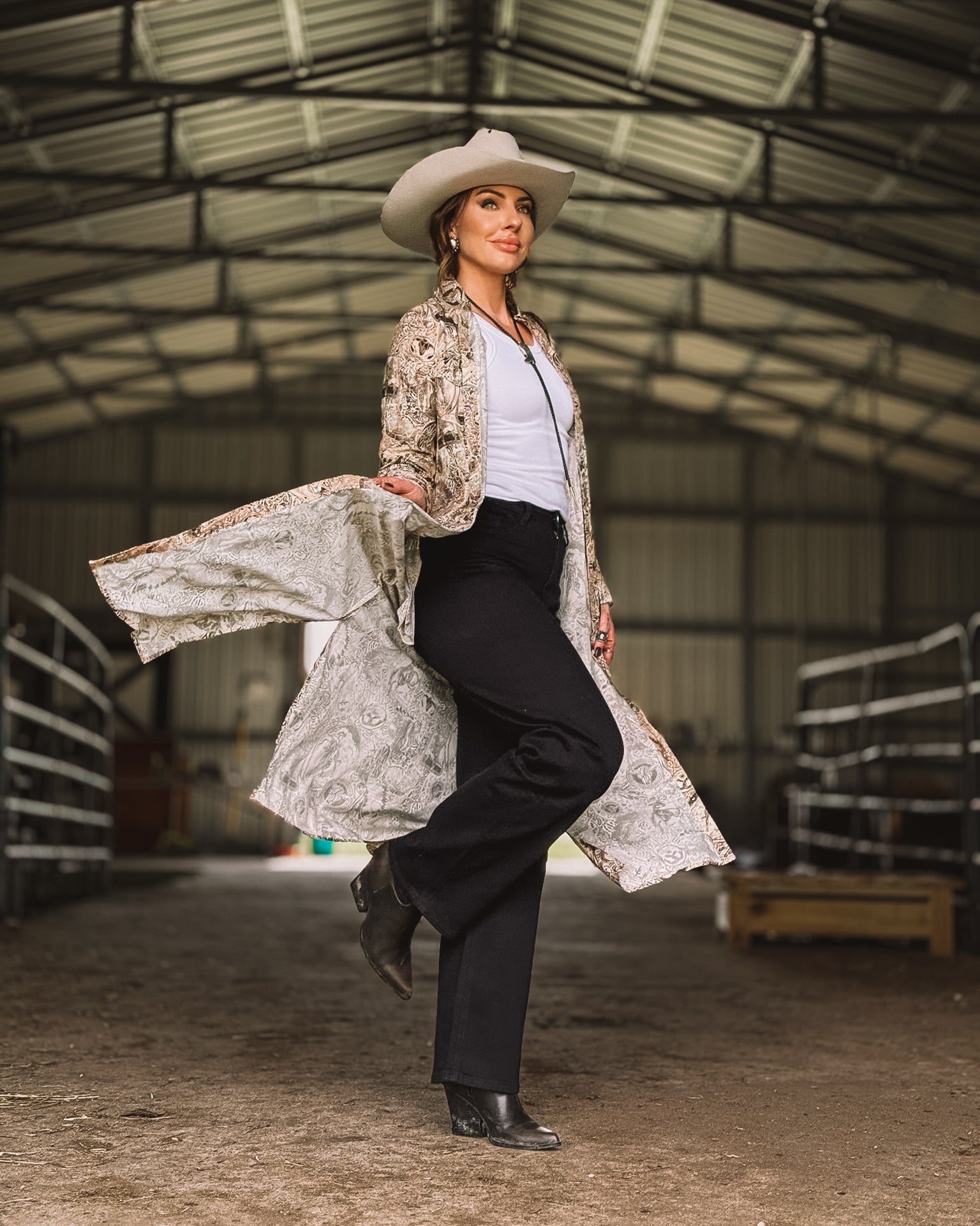 Woman in western boho outfit with cowboy hat, embroidered duster, black pants, and boots in barn