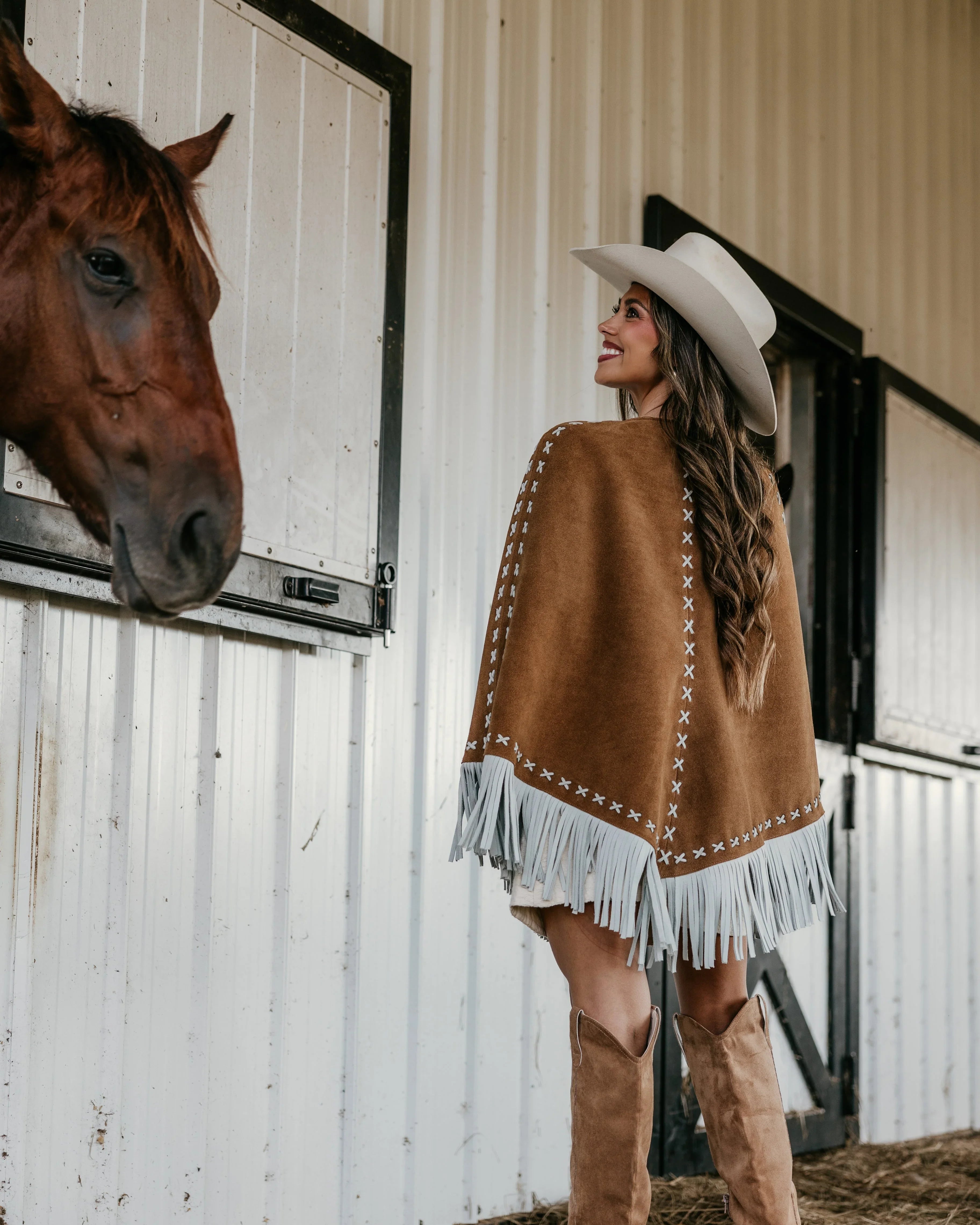 Woman in western fringe poncho, cowboy hat, and boots by horse barn, cowgirl outfit style