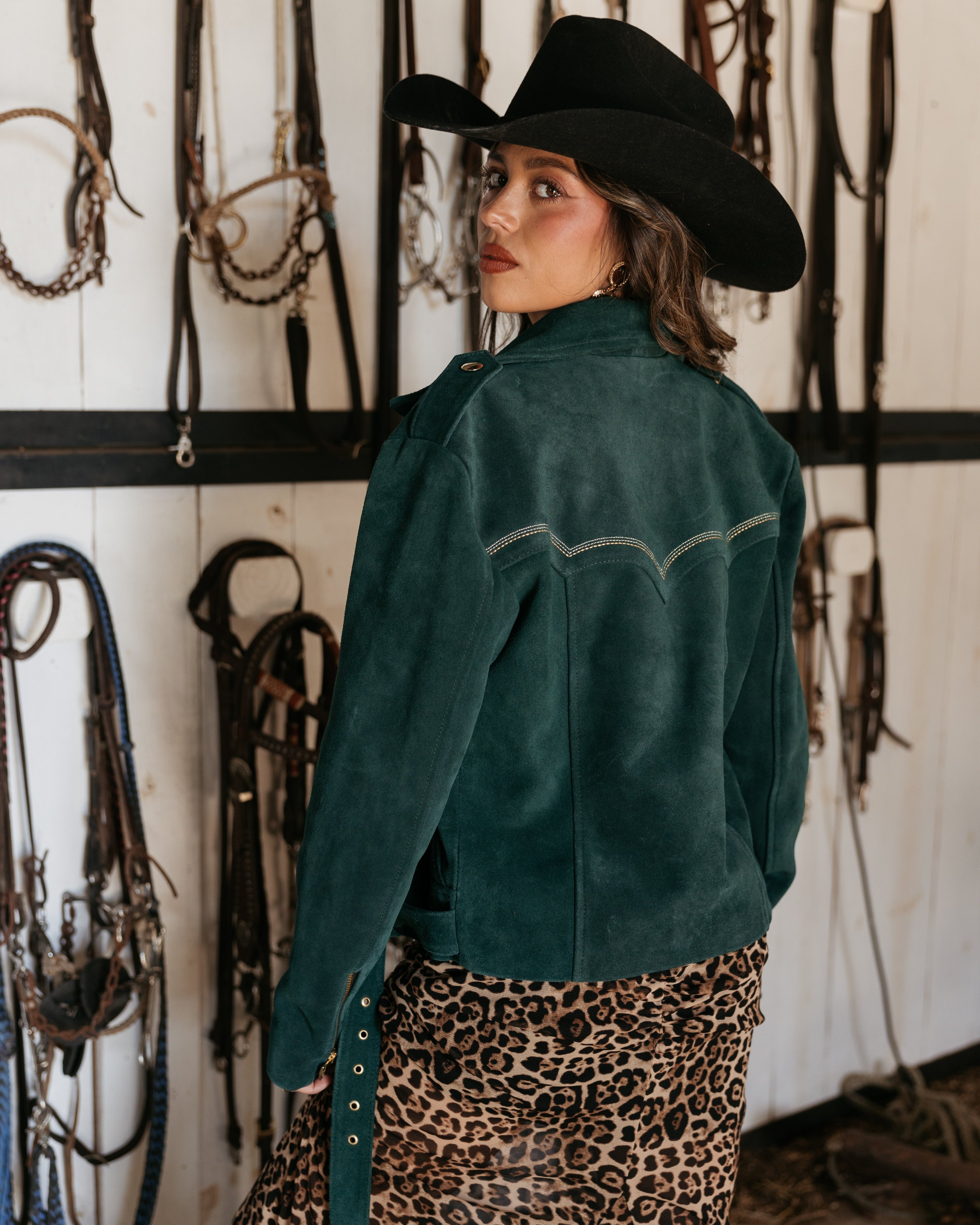 Woman in a suede western jacket, black cowboy hat, and leopard print skirt in a tack room
