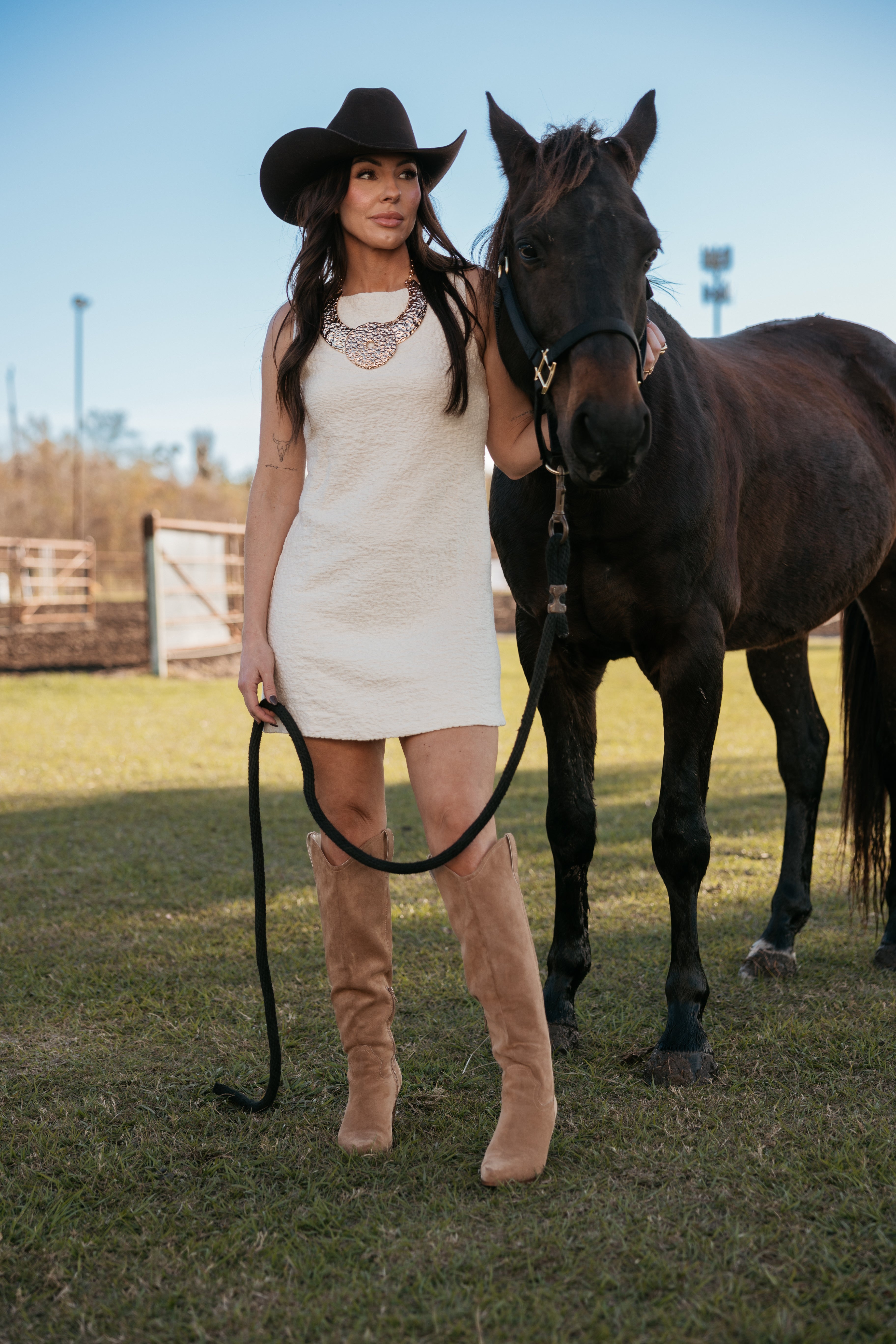 Woman in white western dress, cowboy hat, and boots standing next to a black horse outdoors