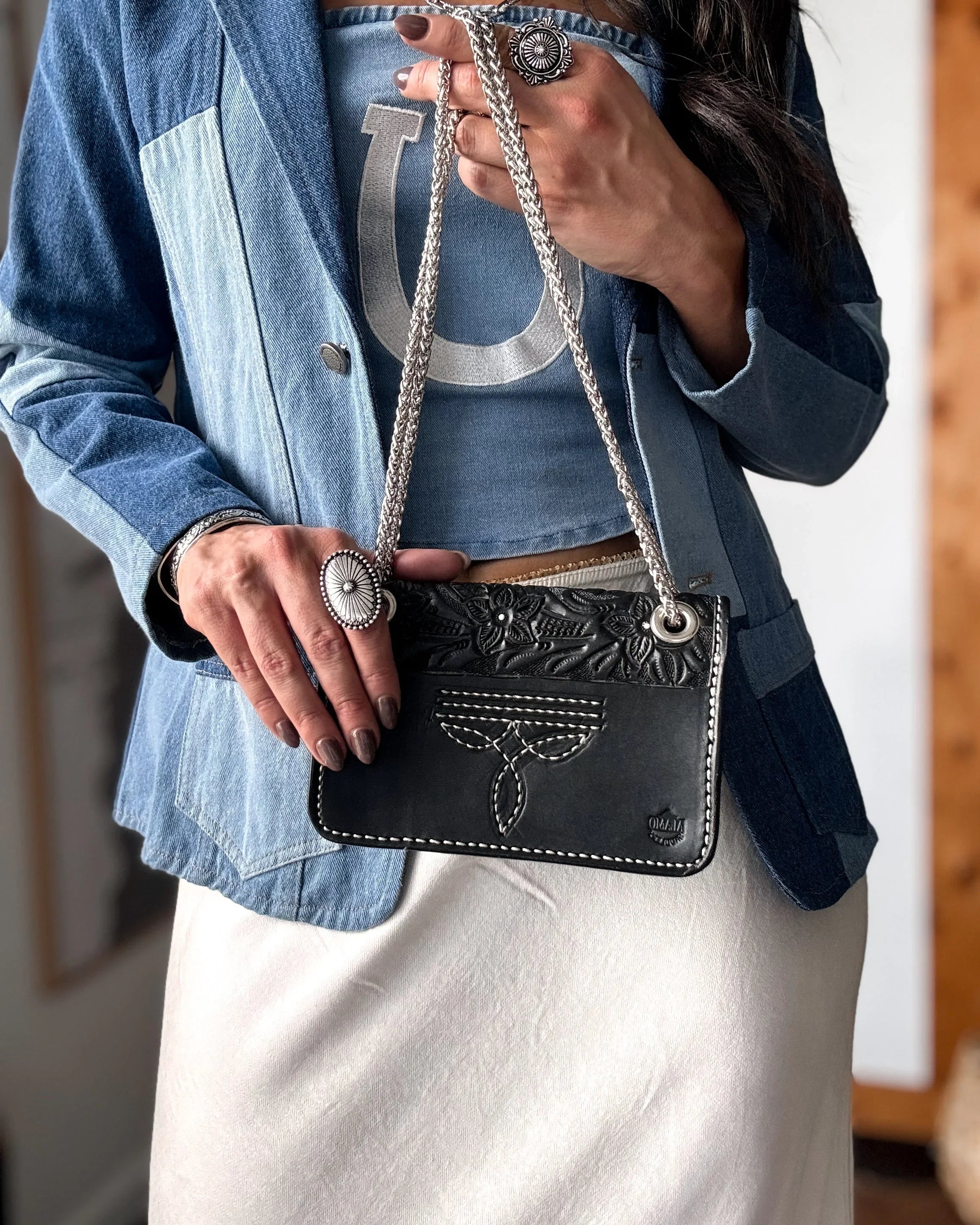 Person holding a black cowhide handbag with intricate designs, wearing a blue denim jacket and white pants.