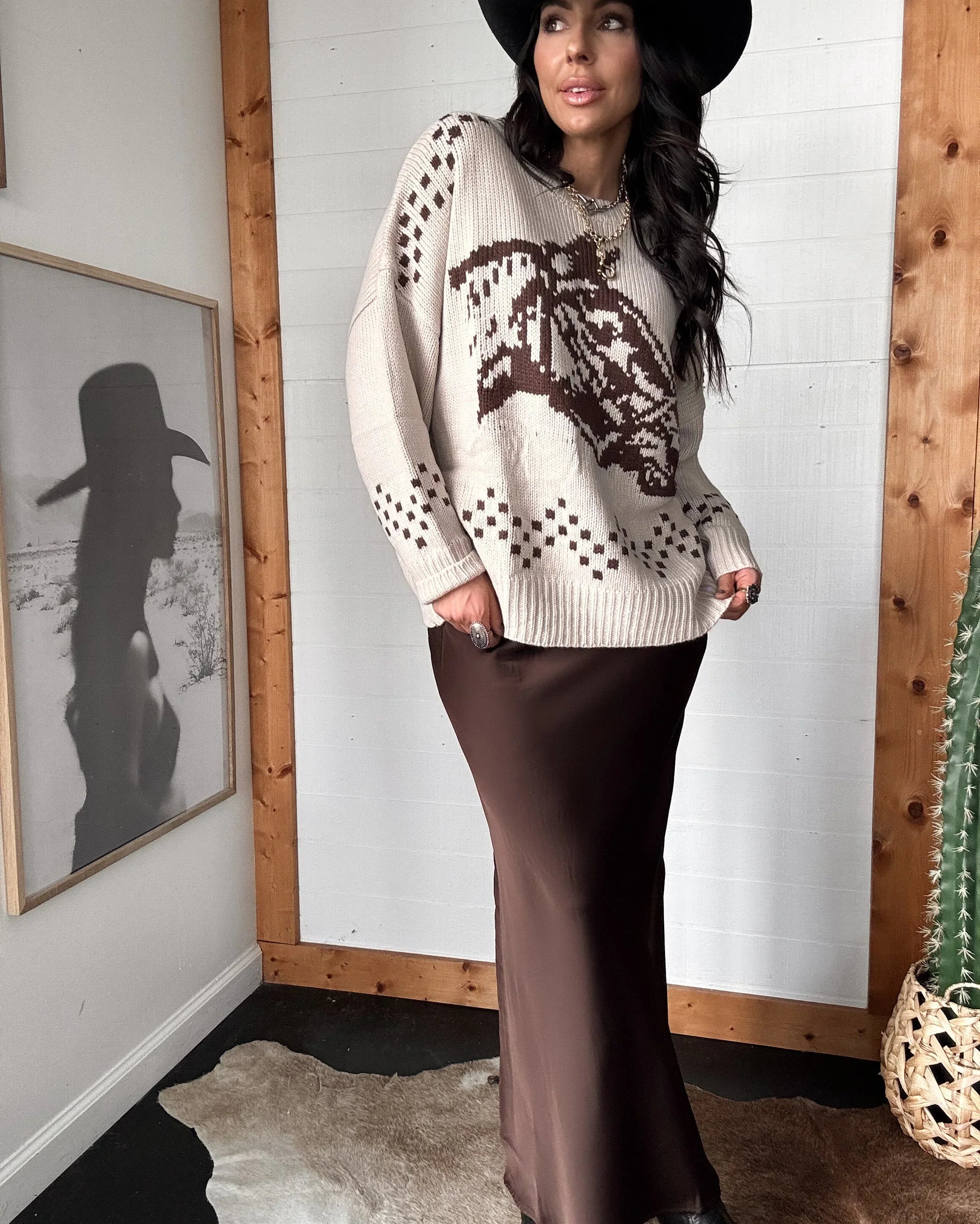 Woman in espresso satin maxi skirt, western graphic sweater, and black cowboy hat, standing indoors.