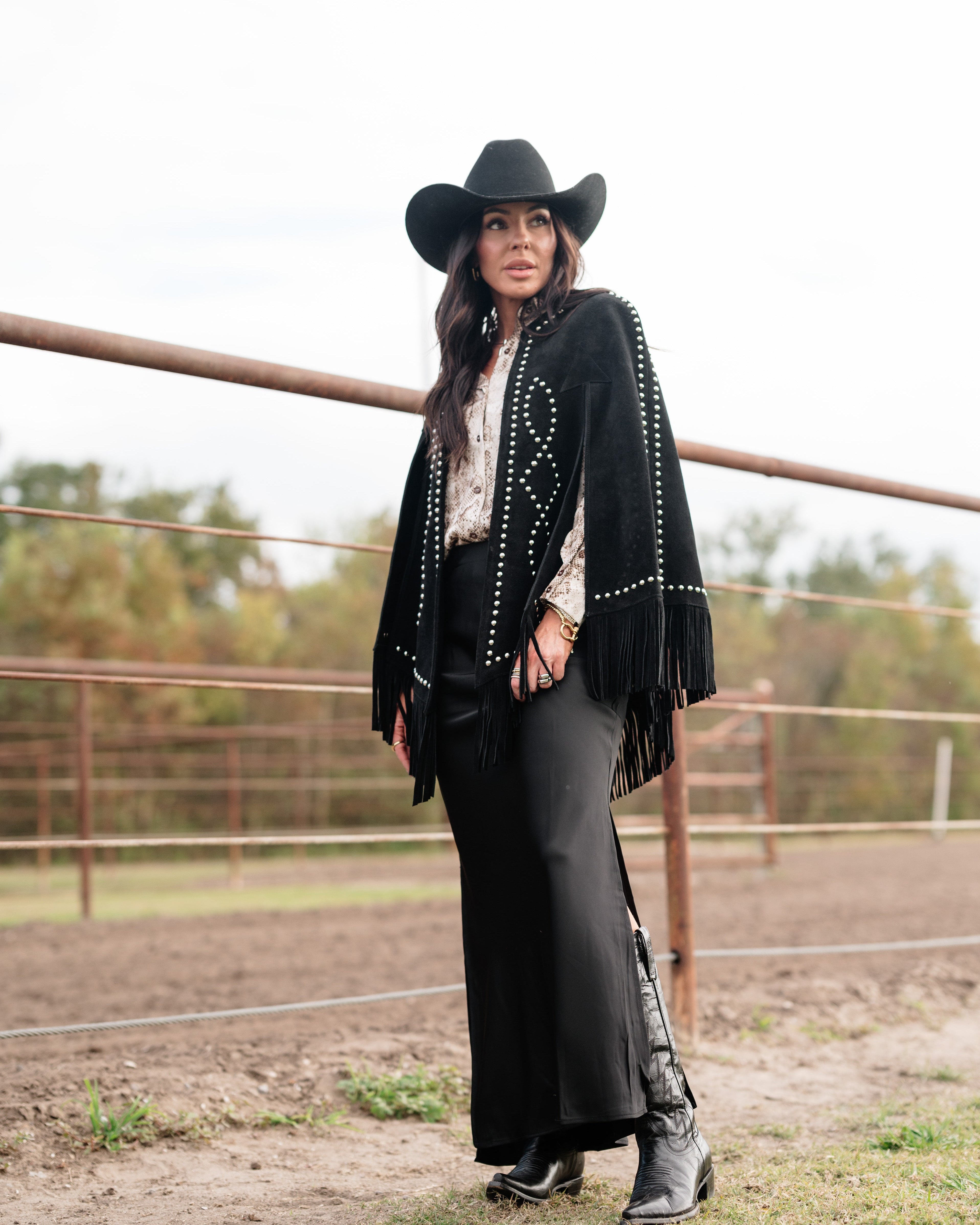 Woman in black cowgirl hat, fringe jacket, maxi skirt, and boots in a western ranch setting