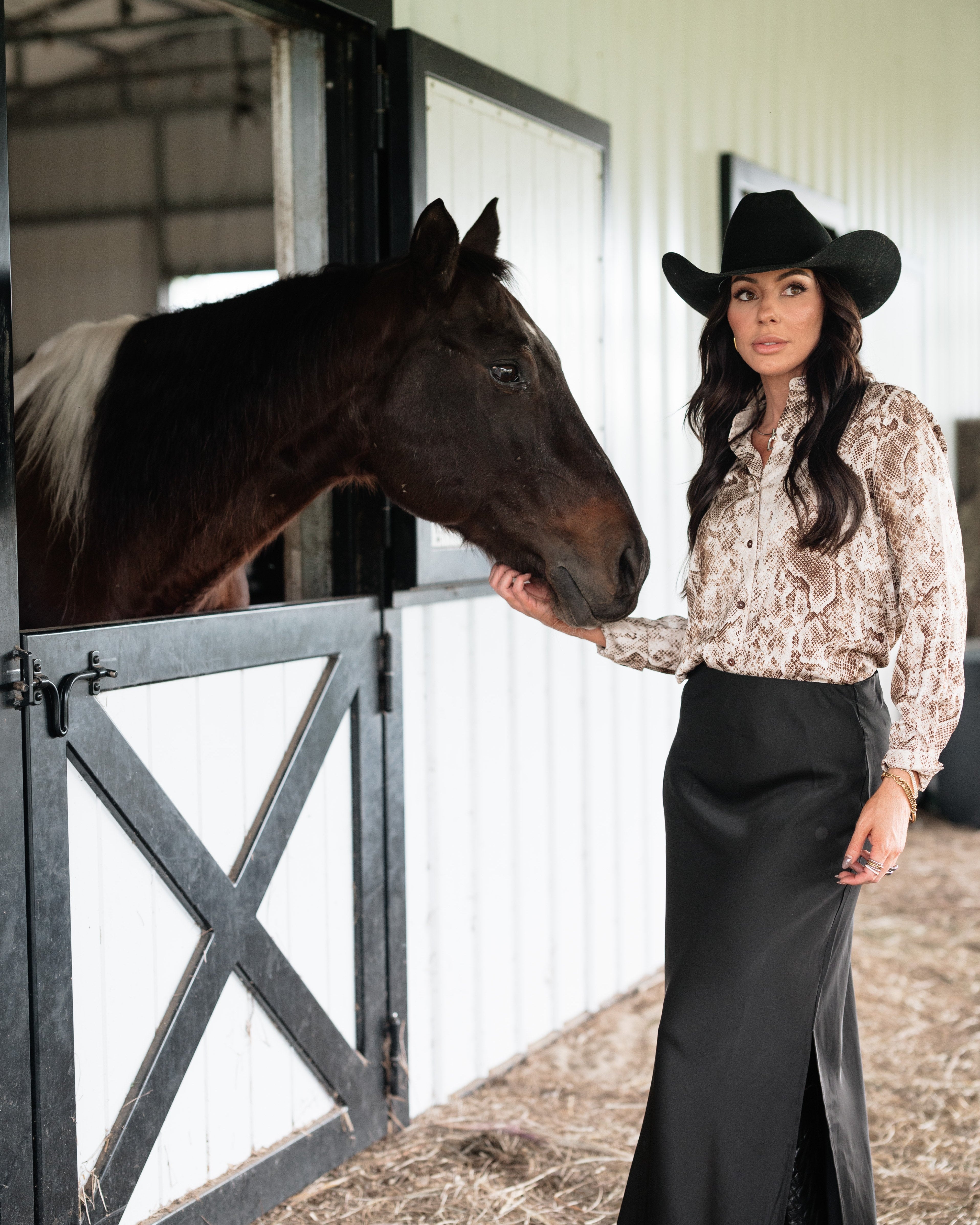 Woman in western cowgirl outfit with black hat and long skirt petting horse by barn