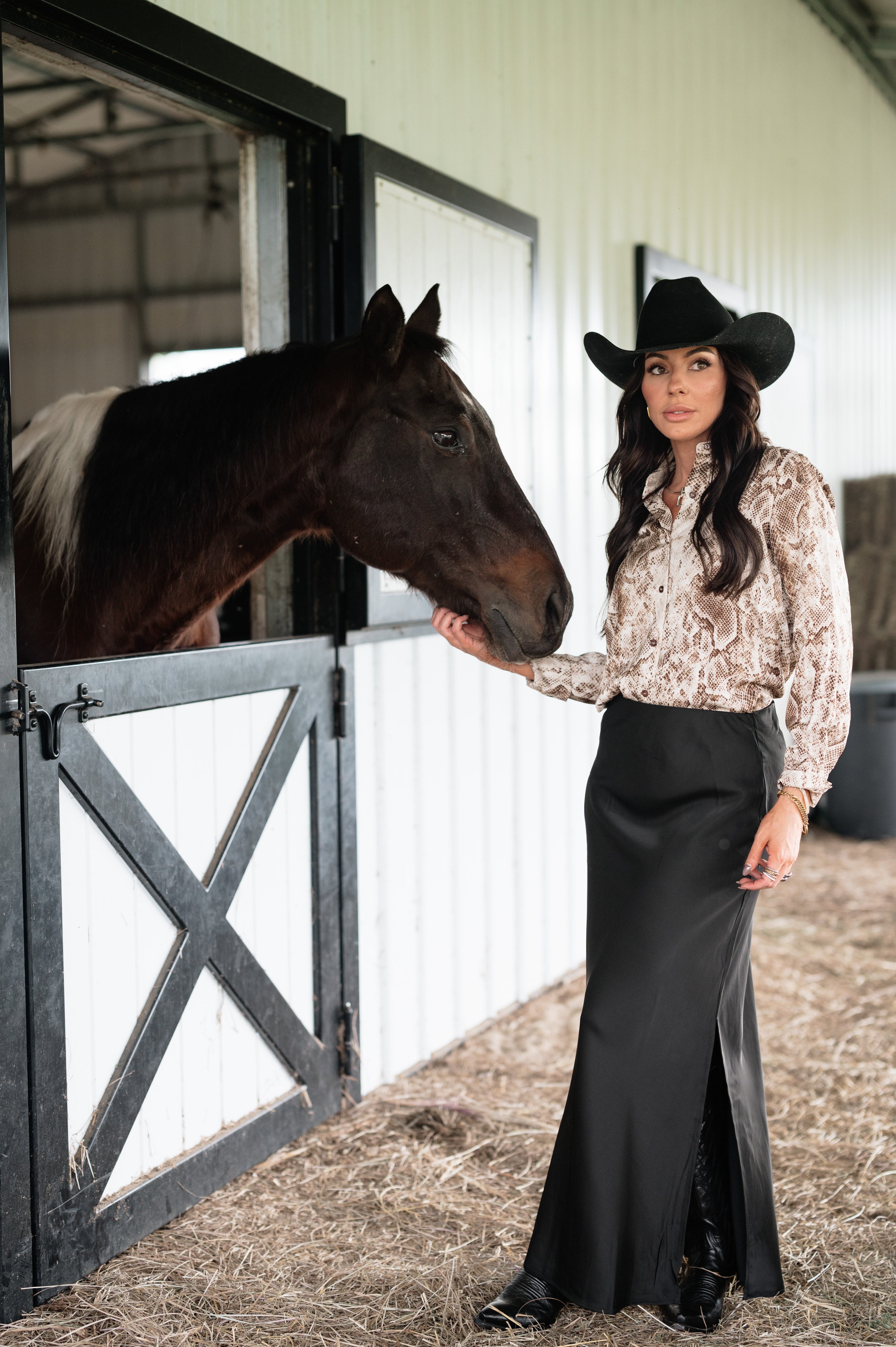 Woman in western cowgirl outfit with black hat and long skirt petting horse by barn