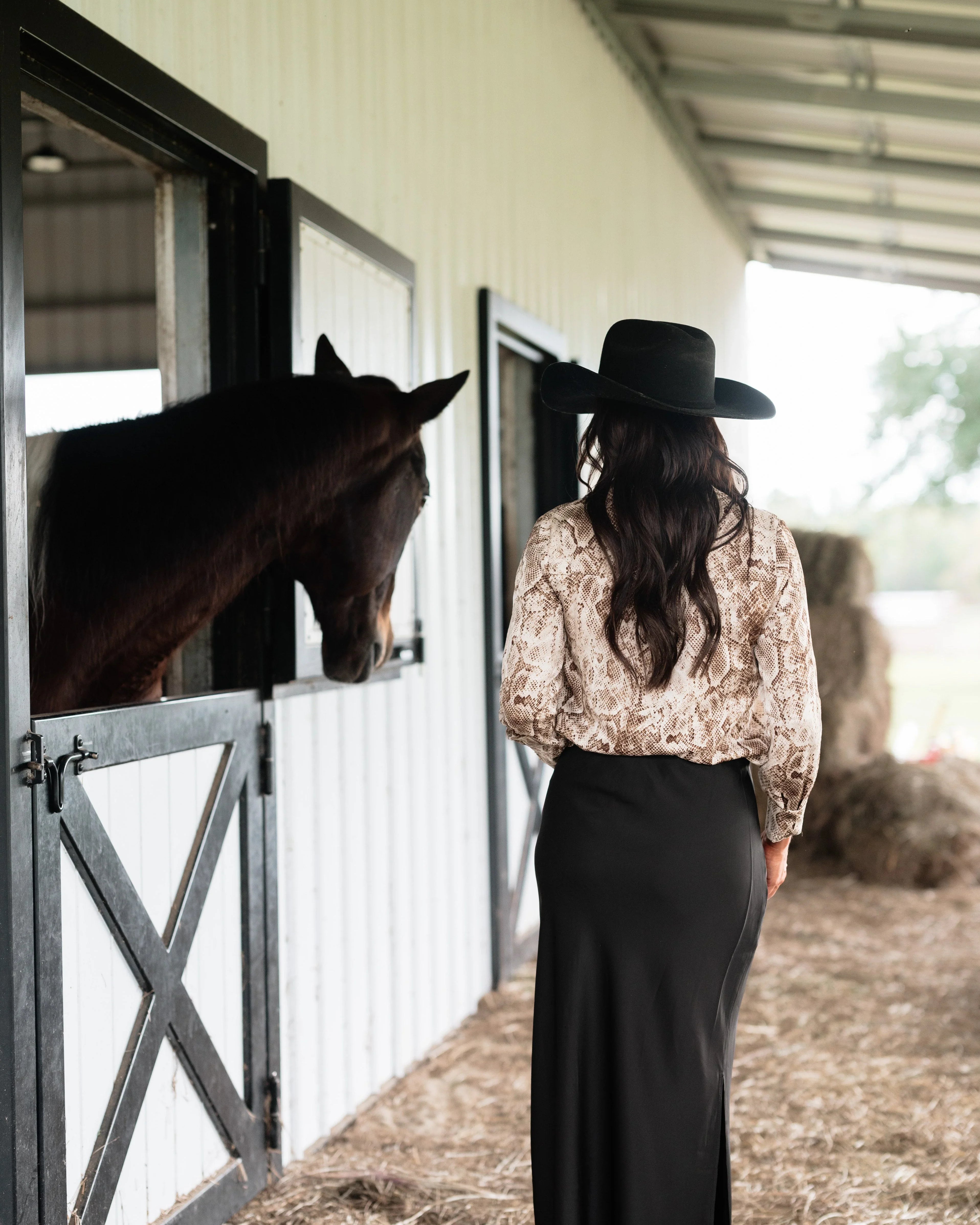 Woman in snakeskin blouse, black maxi skirt, and black hat standing by horse stable