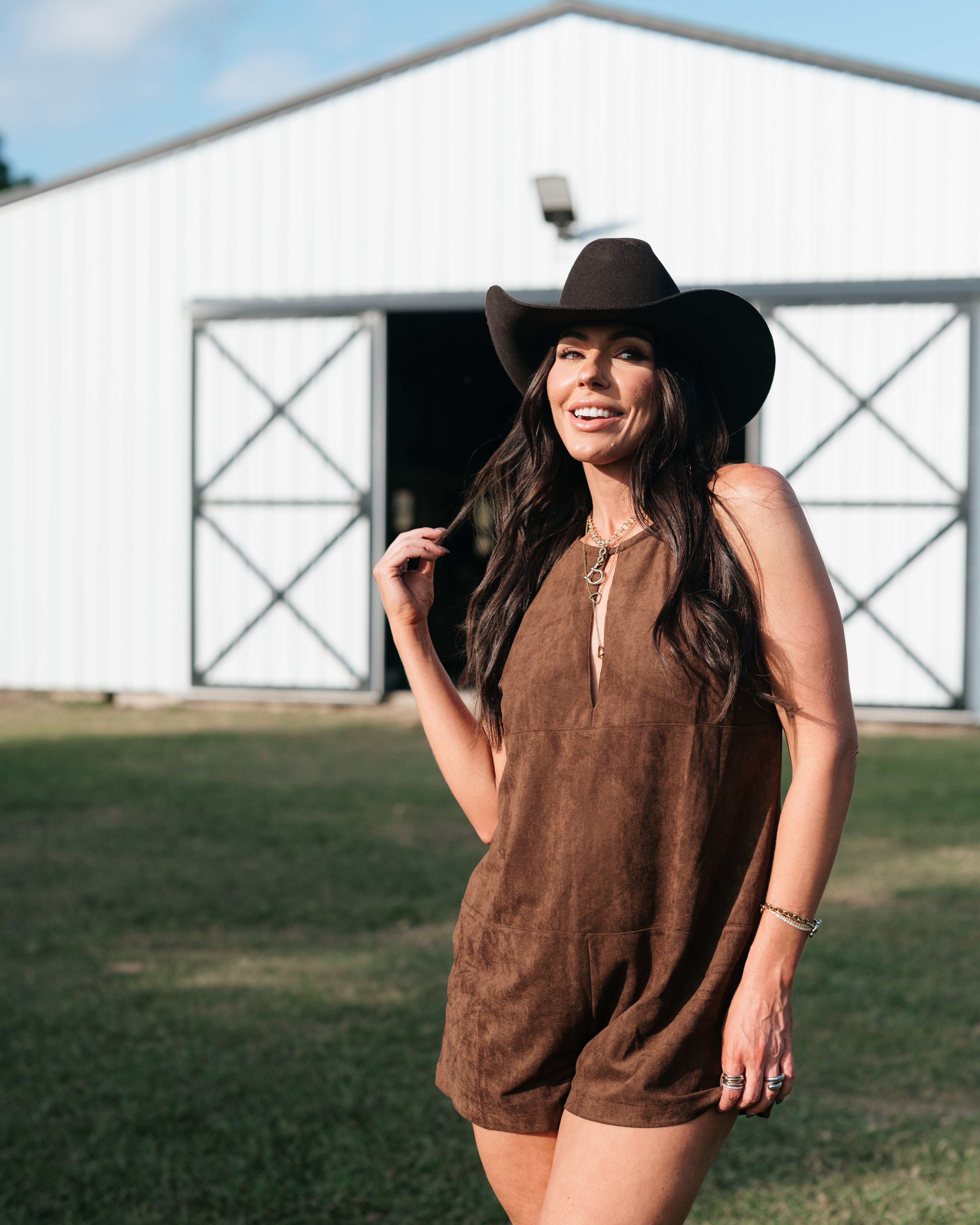 Woman in brown suede romper and black cowboy hat posing outside a barn, western fashion style