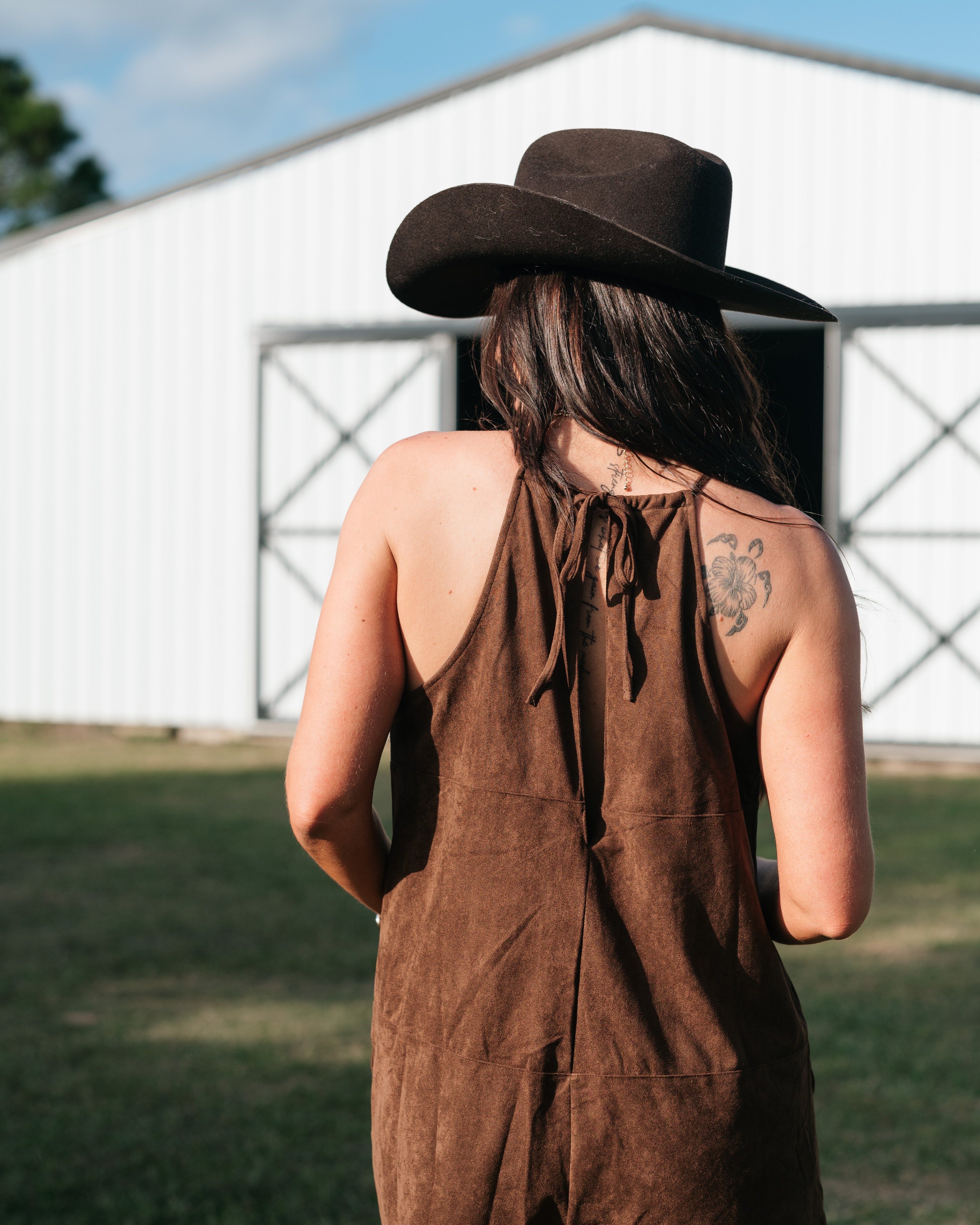 Woman in brown suede romper and black cowboy hat outdoors near a barn, western boho style