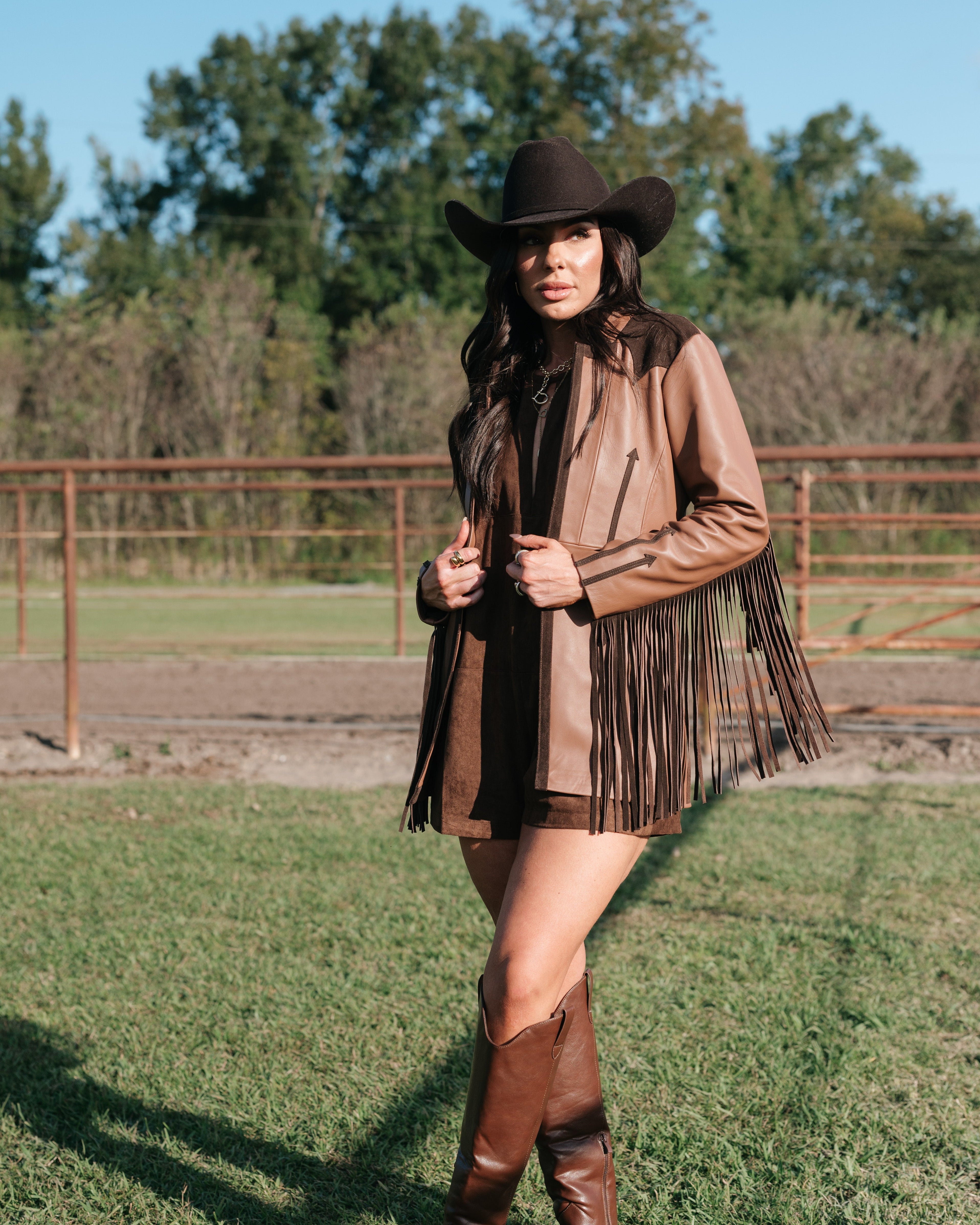 Woman in brown fringe western jacket, cowboy hat, and tall boots standing outdoors