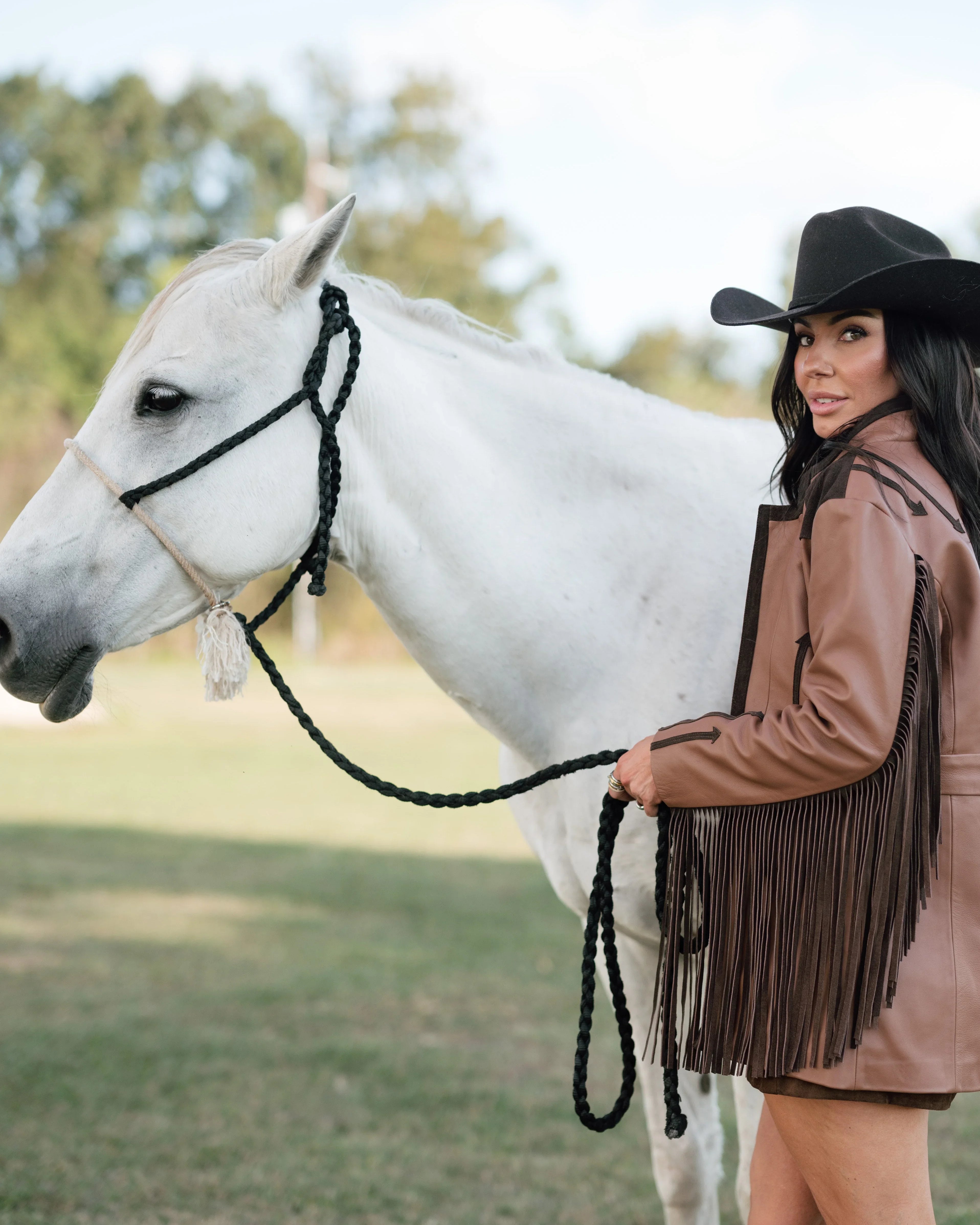Woman in fringe western jacket, cowboy hat, and boots holding white horse outdoors