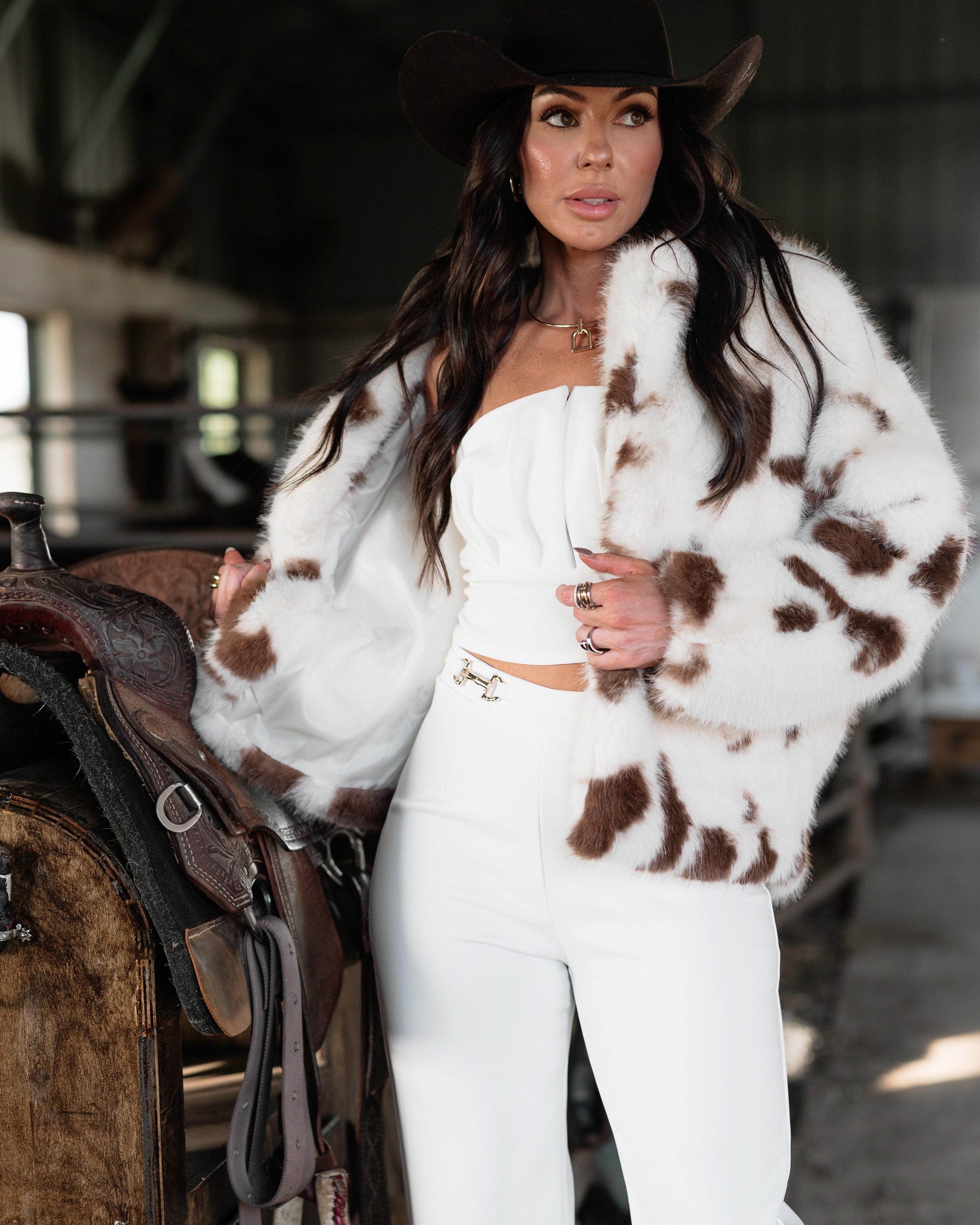 Woman in cow print faux fur jacket, white western outfit, and black cowboy hat by a saddle