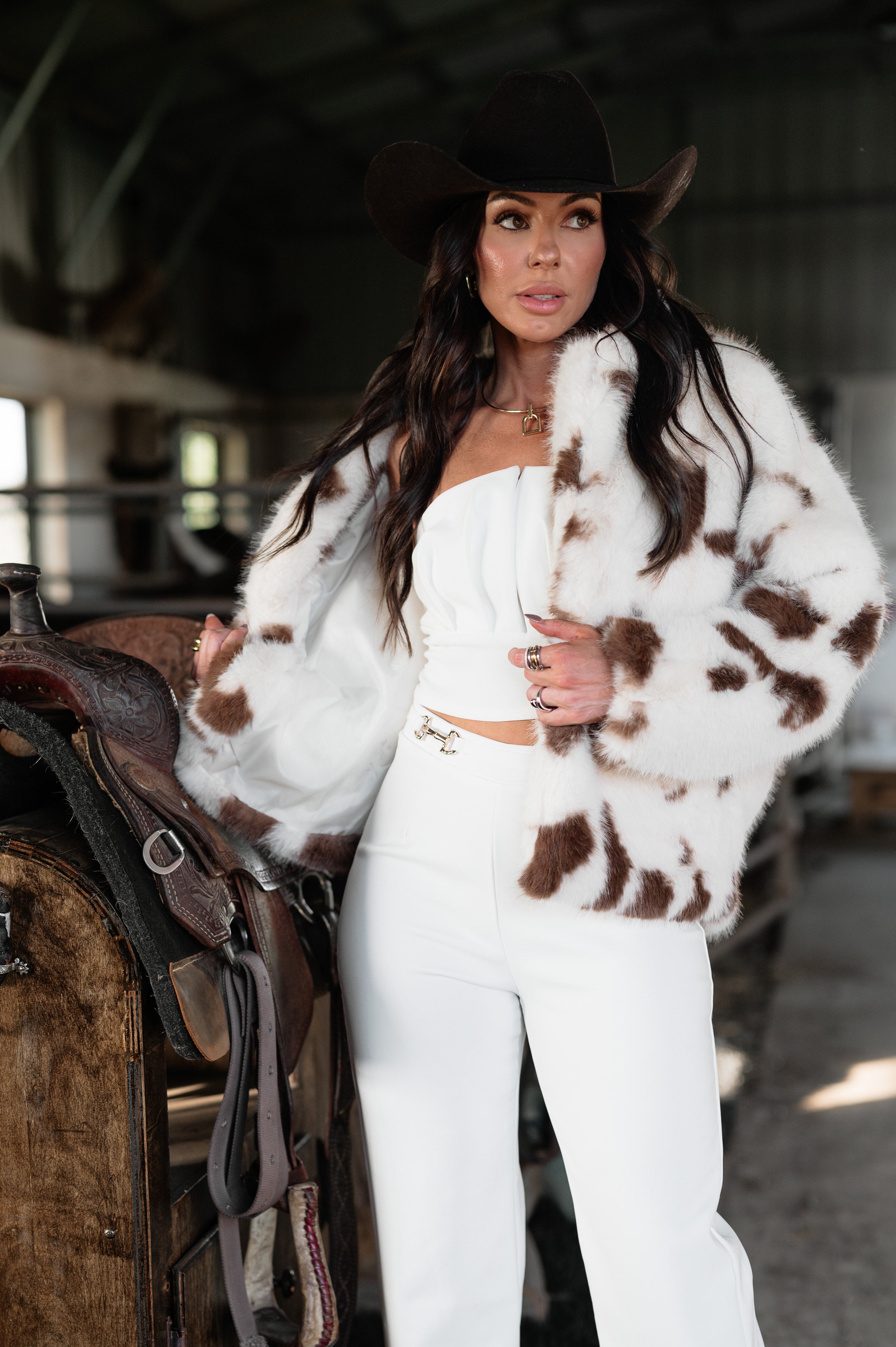 Woman in cow print faux fur jacket, white western outfit, and black cowboy hat by a saddle