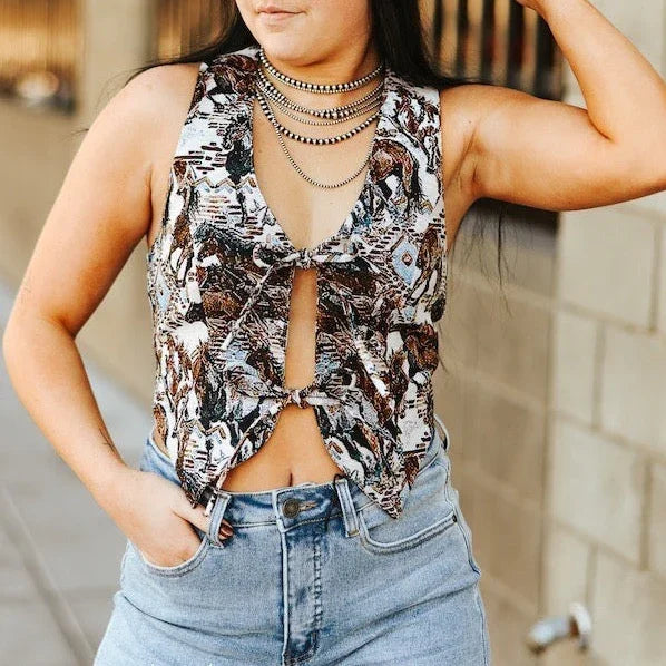 Woman wearing a black cowboy hat, Western horse jacquard halter top with geometric pattern ,and jeans against a brick wall.
