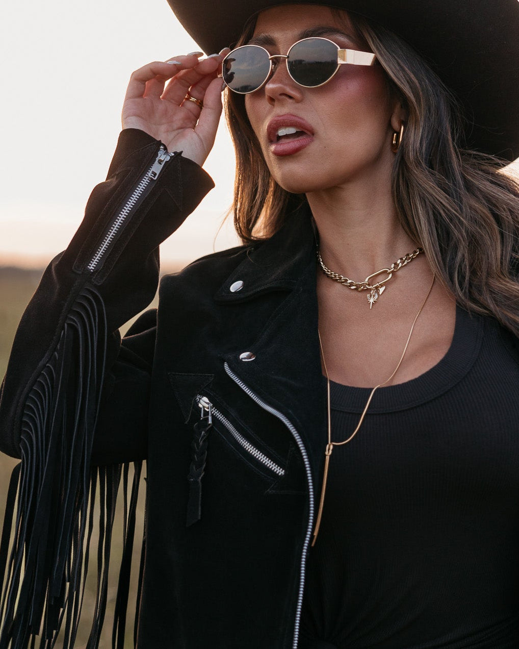 Woman in a black fringe jacket, black tank top, and gold oval framed glasses standing in a field.