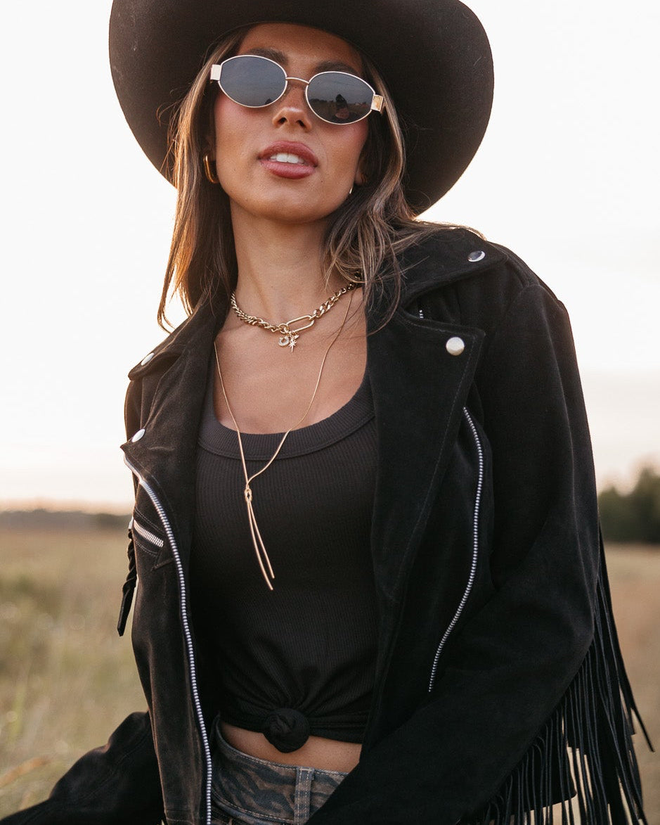 Woman in a black fringe jacket, black tank top, and gold oval framed glasses standing in a field.