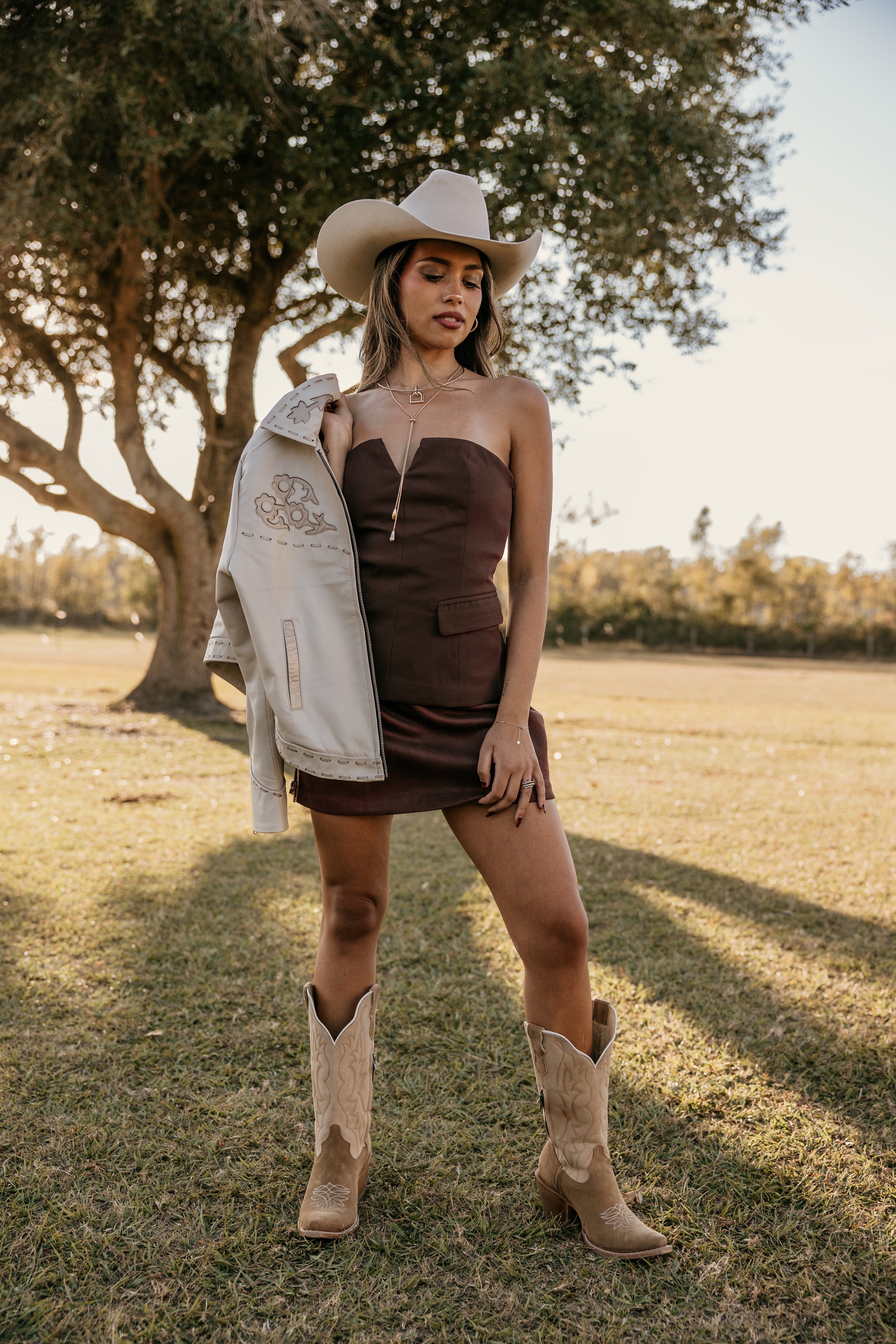 Woman in strapless brown western dress, cowboy boots, and hat holding embroidered jacket outdoors