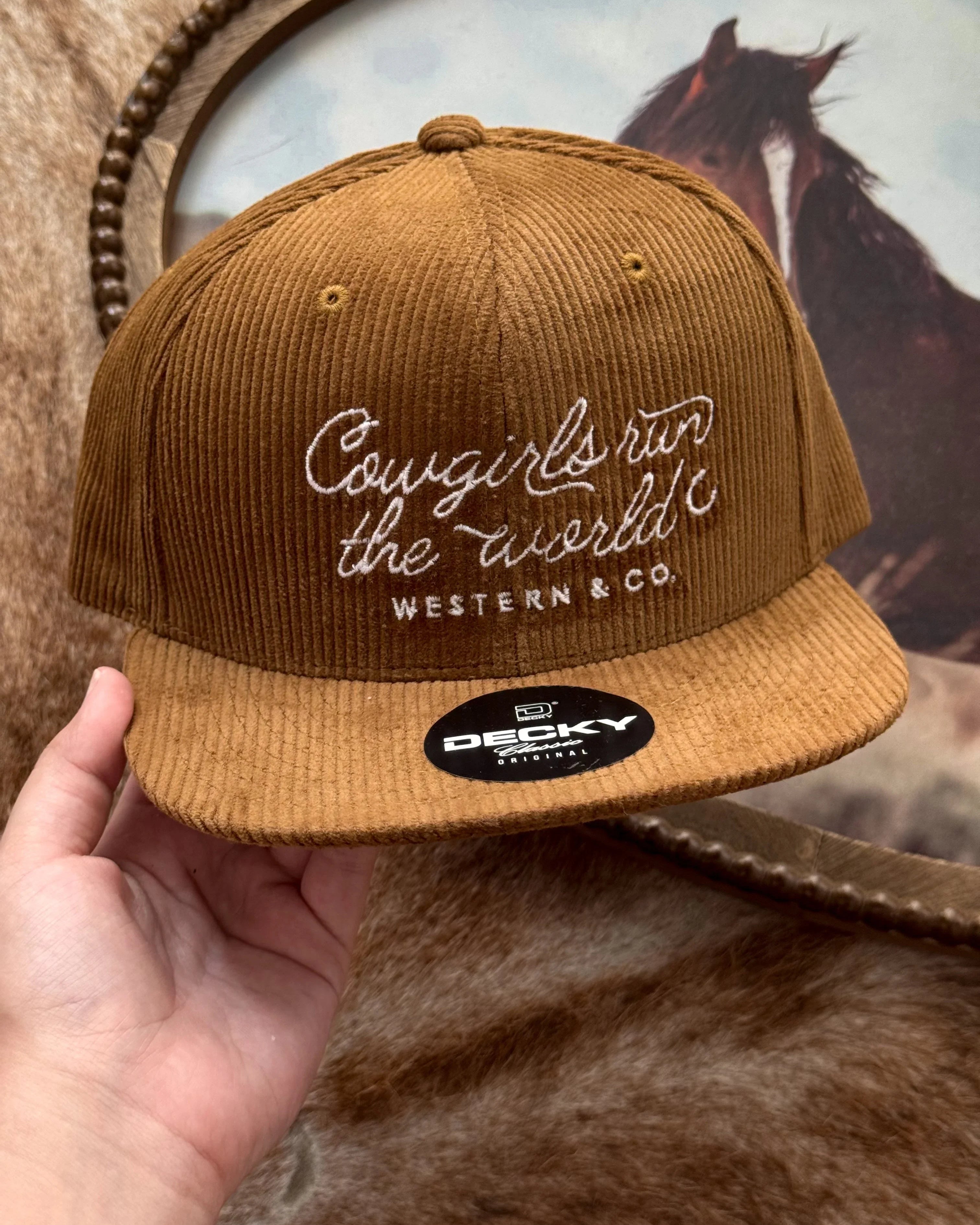 Brown corduroy western baseball hat with 'Cowgirls run the world' embroidery, held by hand.