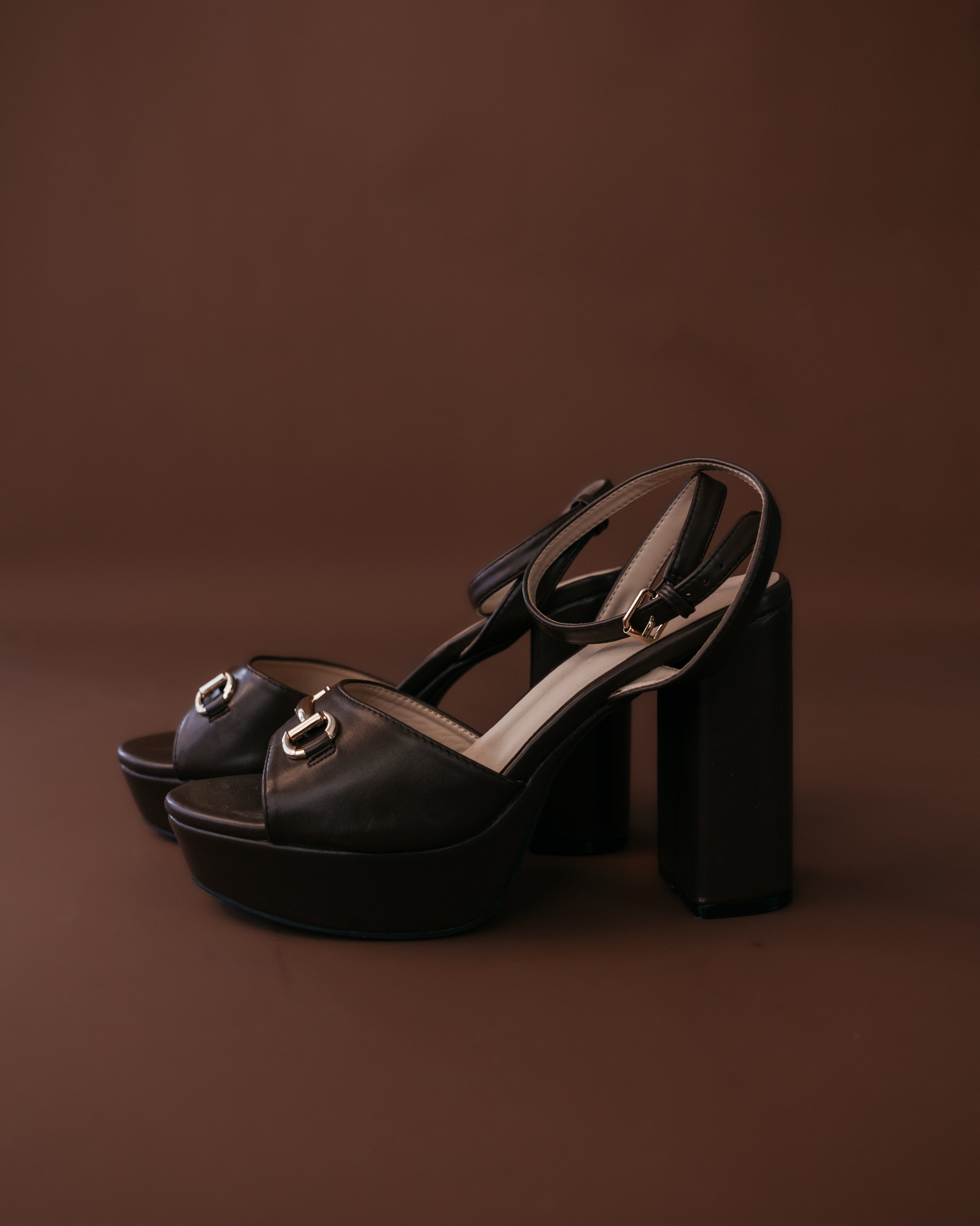 Black platform block heel sandals with gold horsebit detail on brown background